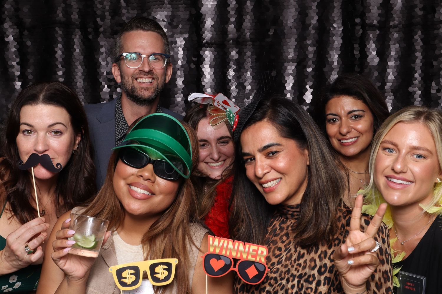 Group of friends posing in a photo booth with fun props including glasses and winner signs against a silver backdrop.