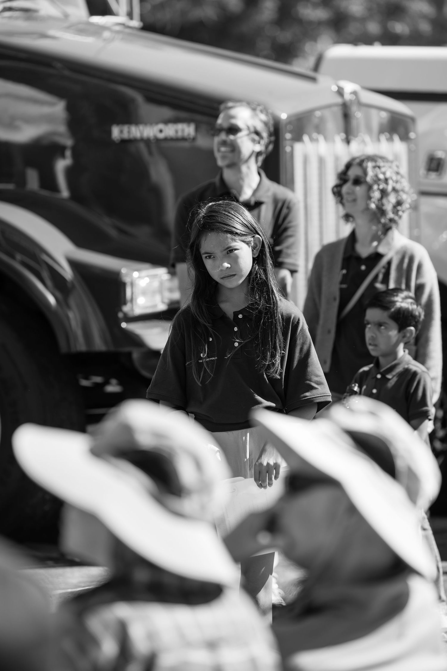 Black and white photo of a young girl with braids standing outdoors near a Kenworth truck with adults behind her.