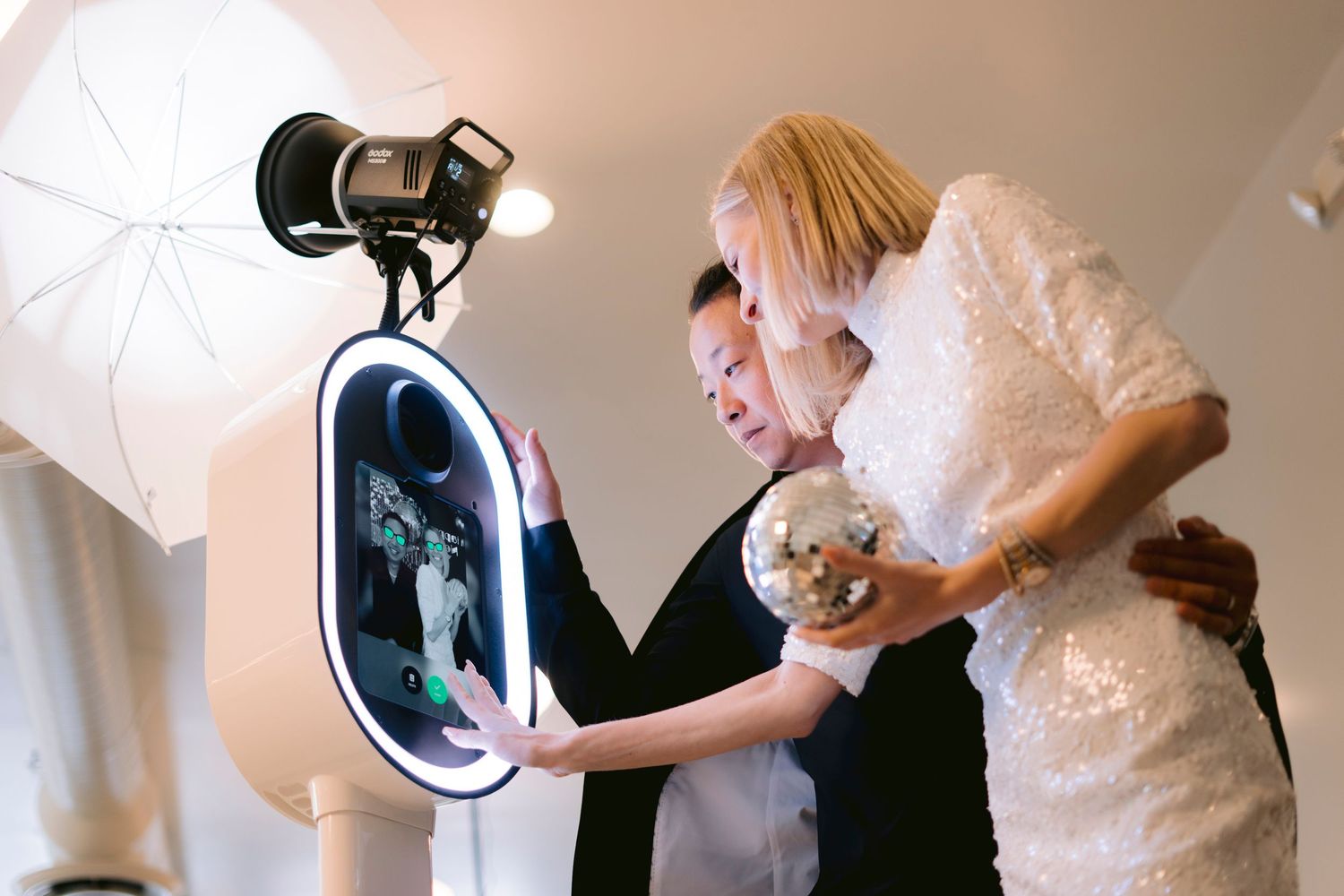 Two guests holding a mini disco ball review a black and white photo on the glowing Lune photo booth screen.