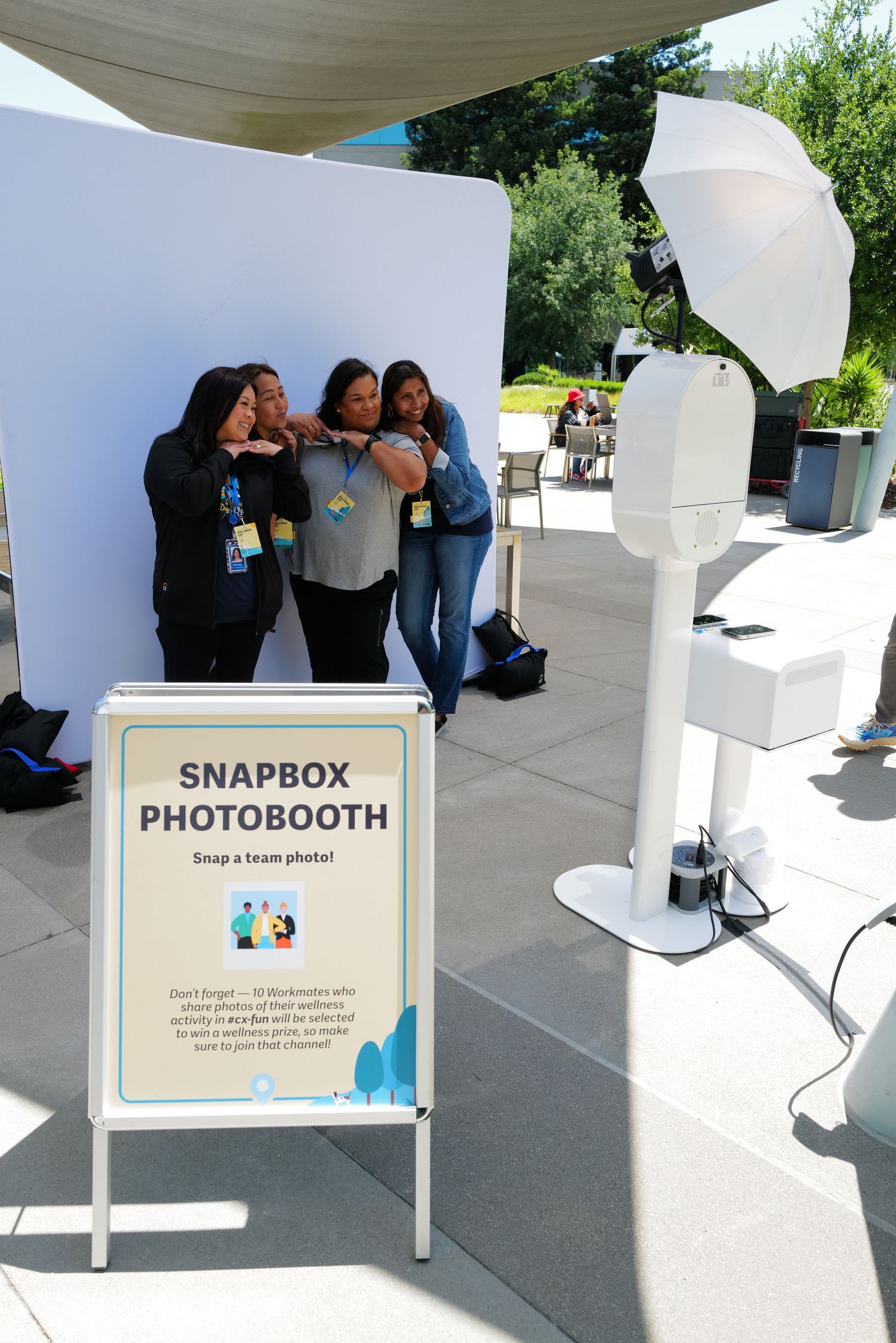 Three women pose playfully at a Snapbox Photo Booth setup outdoors at a corporate event with a white backdrop.