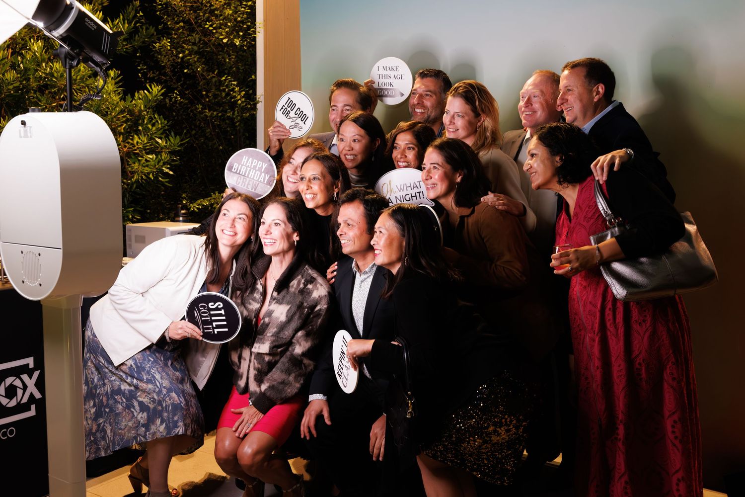 Large group of smiling people posing together at a photo booth event, holding props and signs.