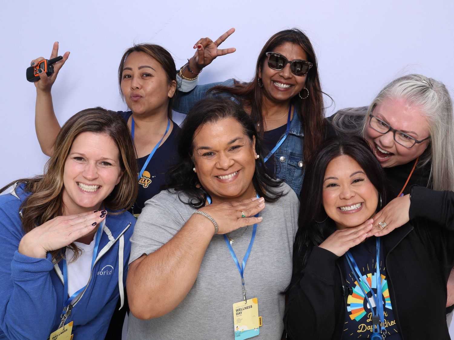 Six women posing playfully at an event, wearing blue lanyards and badges, smiling and making fun gestures.