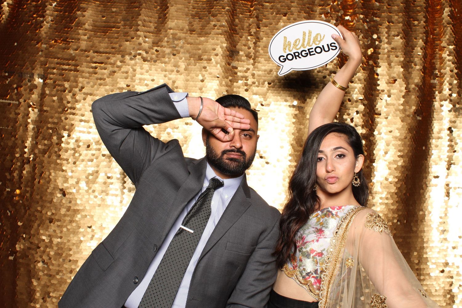 Couple posing playfully at photo booth with hello gorgeous sign, gold backdrop.