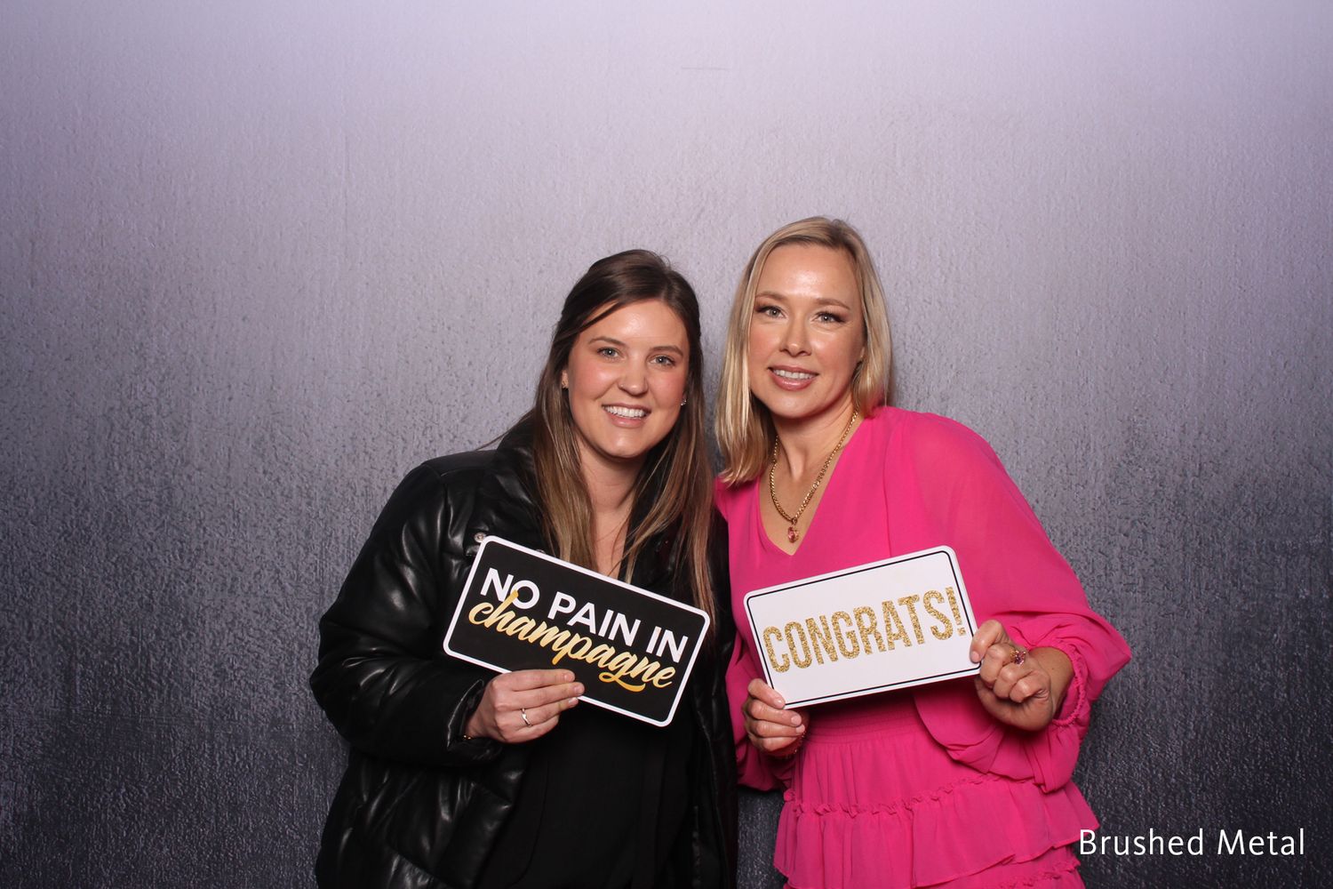 Two women smiling and holding Congrats and No Pain in Champagne signs in photo booth.