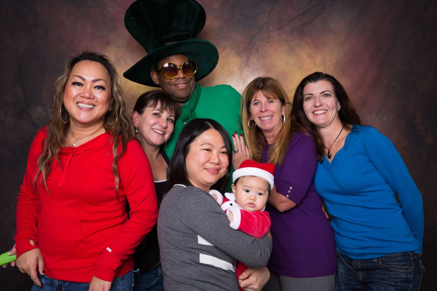 Group of six people posing for a festive photo, including a man in a green hat and a woman holding a baby in a Santa hat. Gensler Holiday