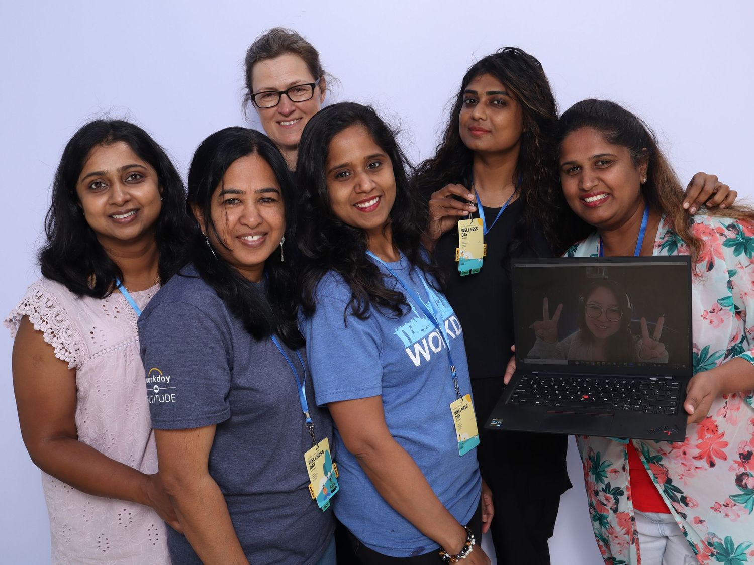 Six women smiling and posing together at a tech event, one holding a laptop displaying a face.