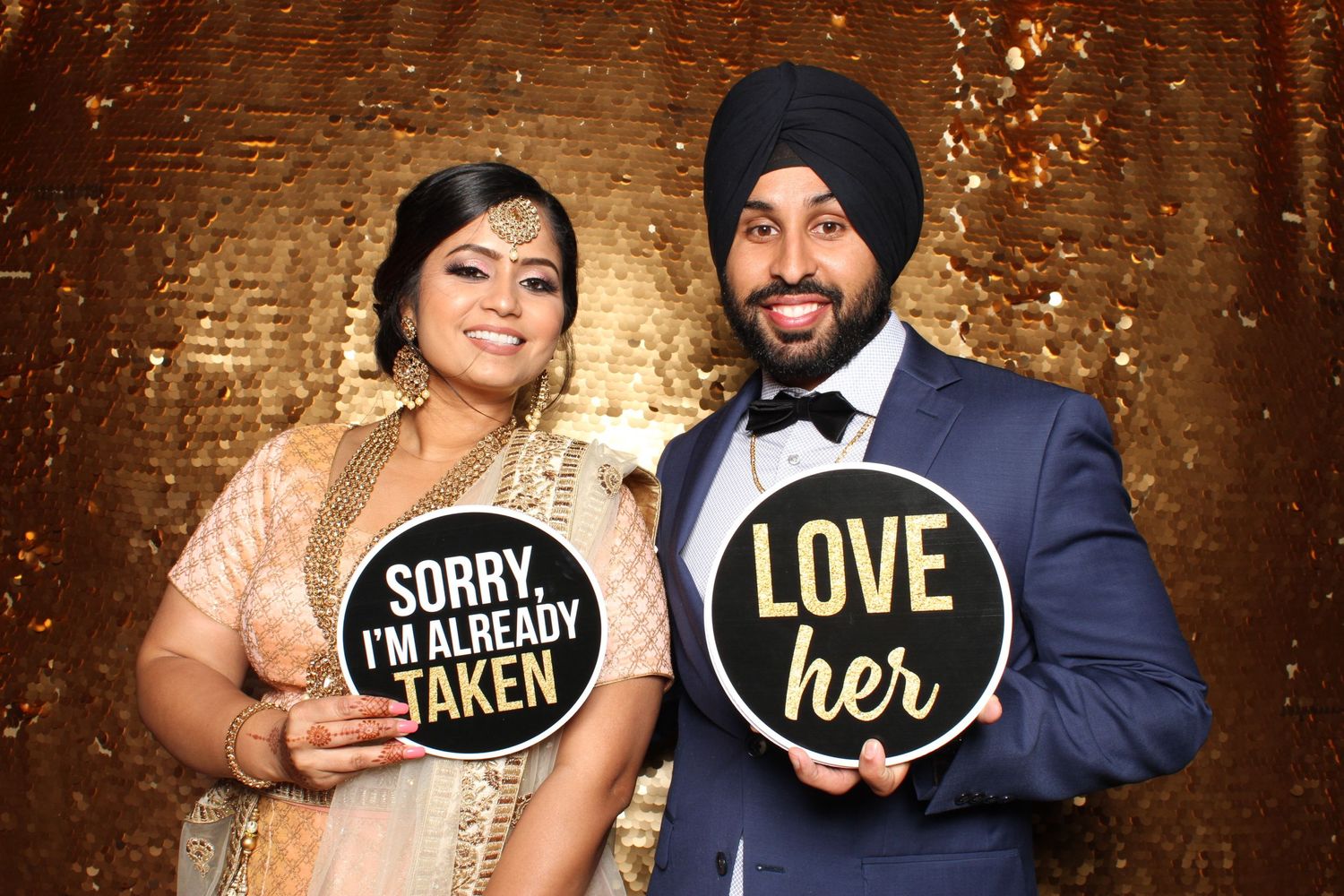 Couple in formal attire holding fun signs at photo booth with gold backdrop.