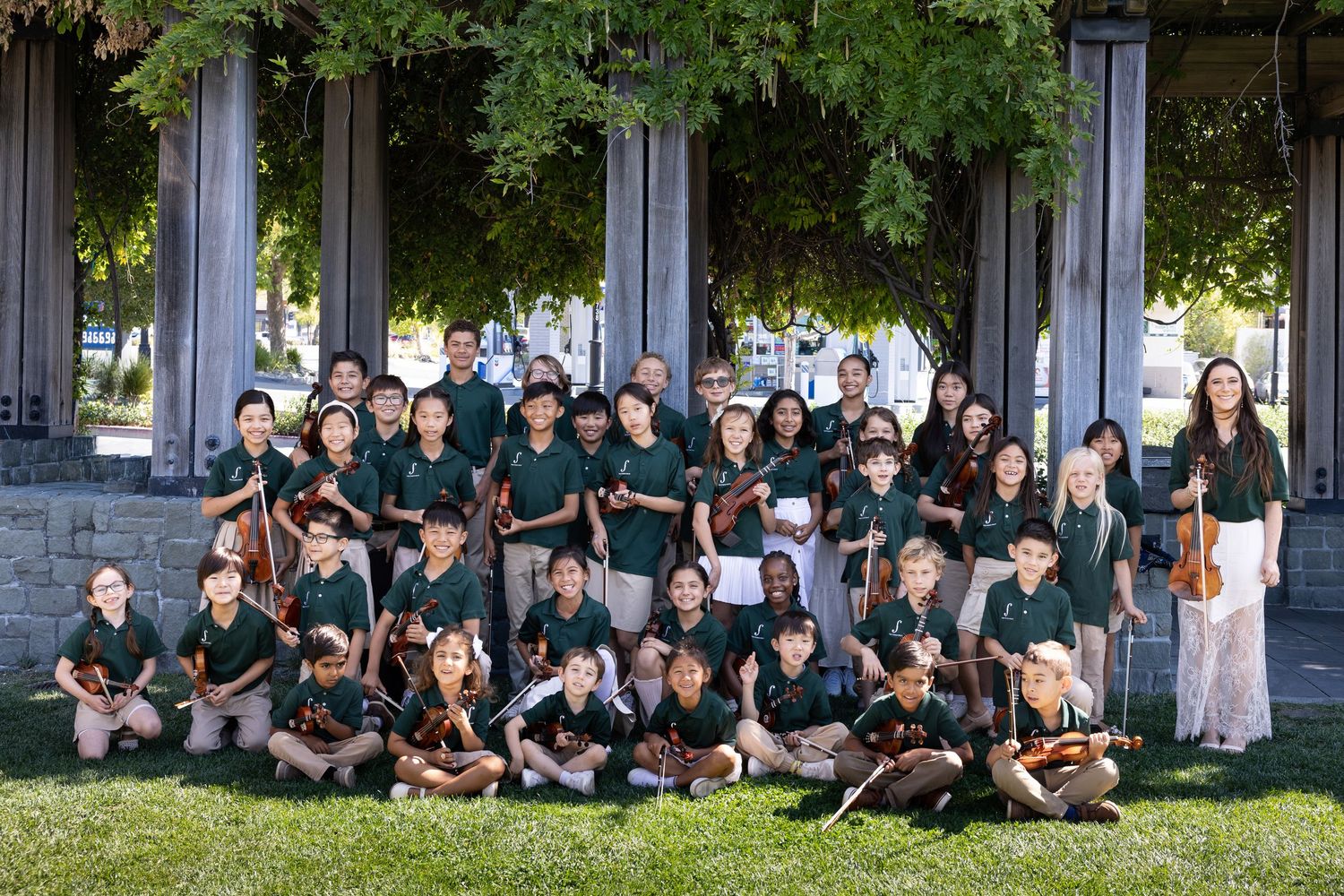Young string orchestra students in green uniforms pose outdoors with violins near stone columns.