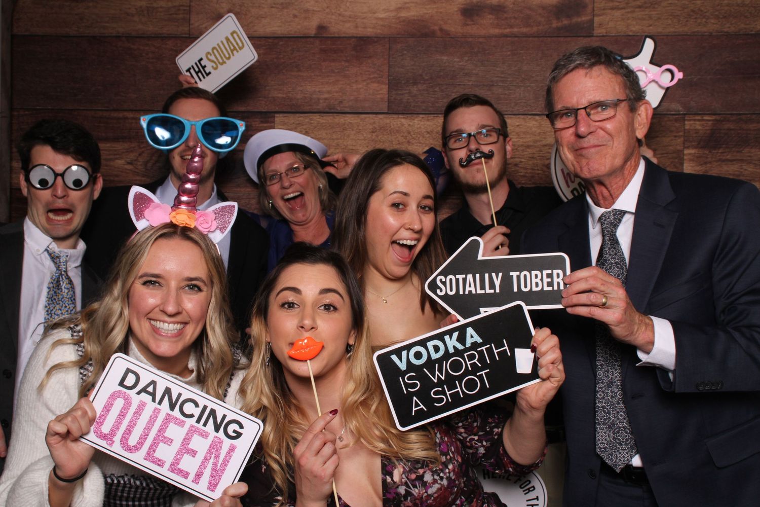 Group of eight party guests pose with fun props and signs at a photo booth.