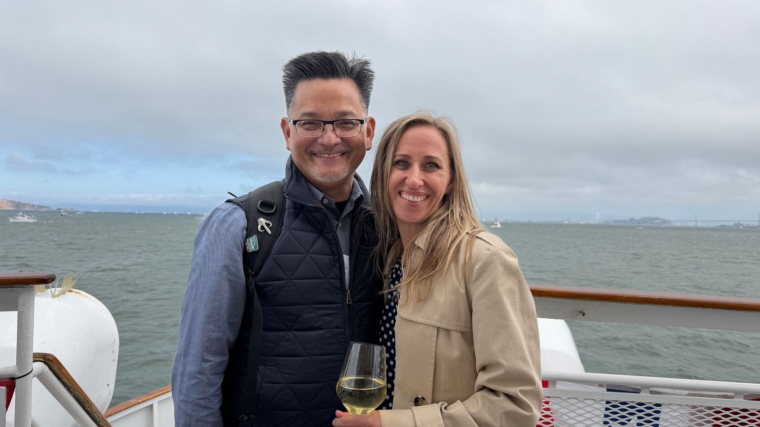 Couple smiling on a boat deck with San Francisco Bay and cloudy skies in the background, woman holding wine glass.