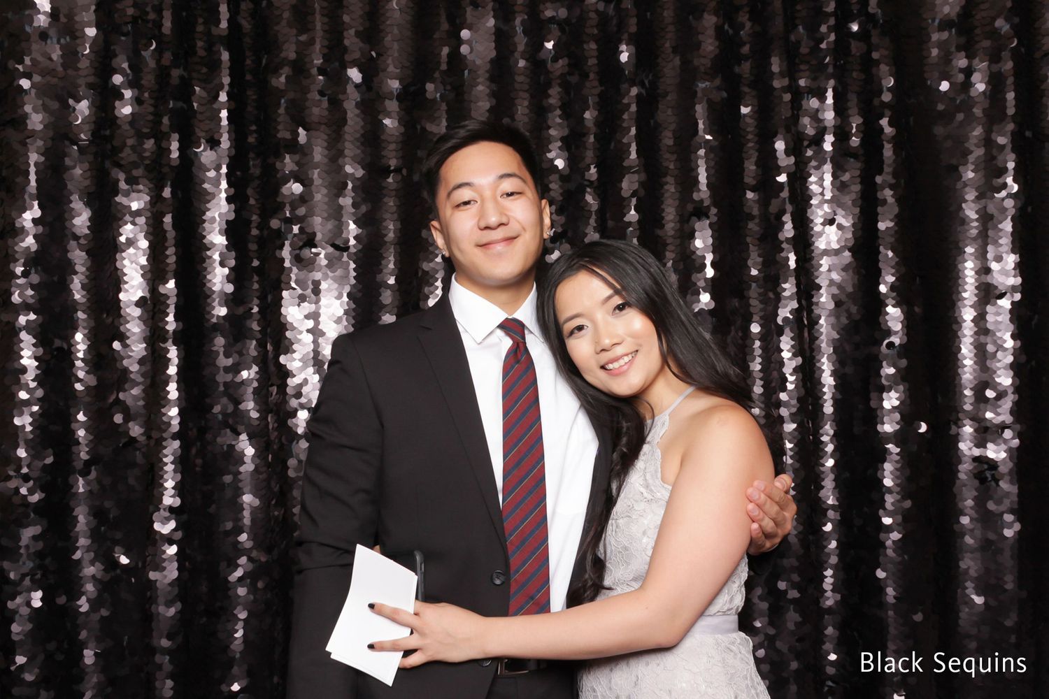 Couple posing together in front of a black sequin backdrop at a formal event, man in suit and woman in white dress.