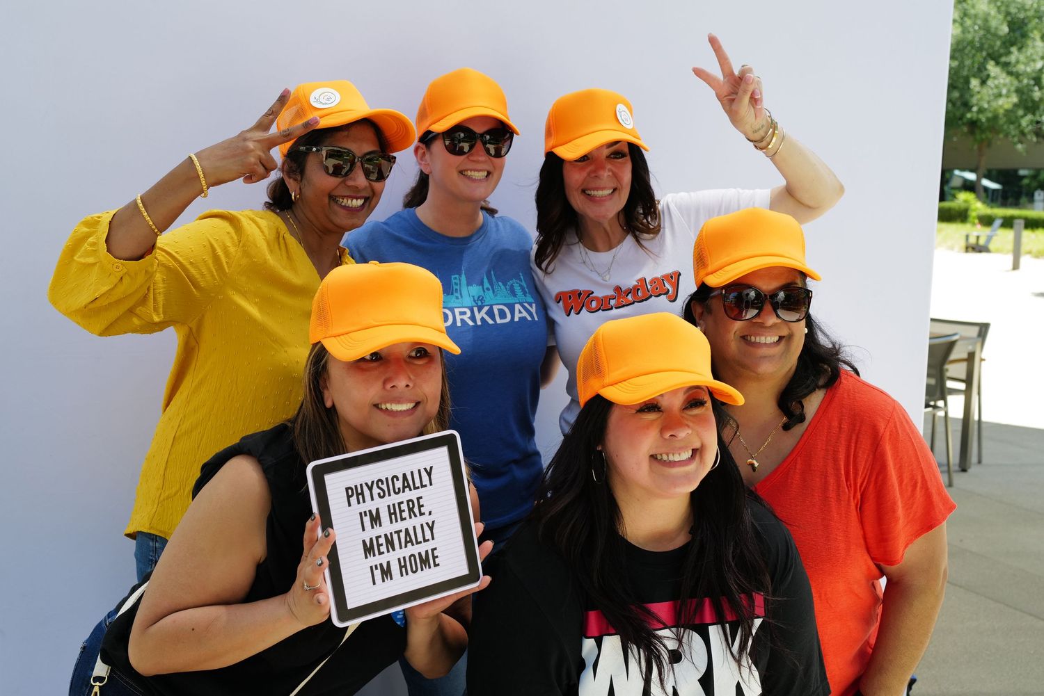 Workday group wearing orange Workday branded hats smiling outdoors, one holding a humorous sign about working remotely. Pleasanton.