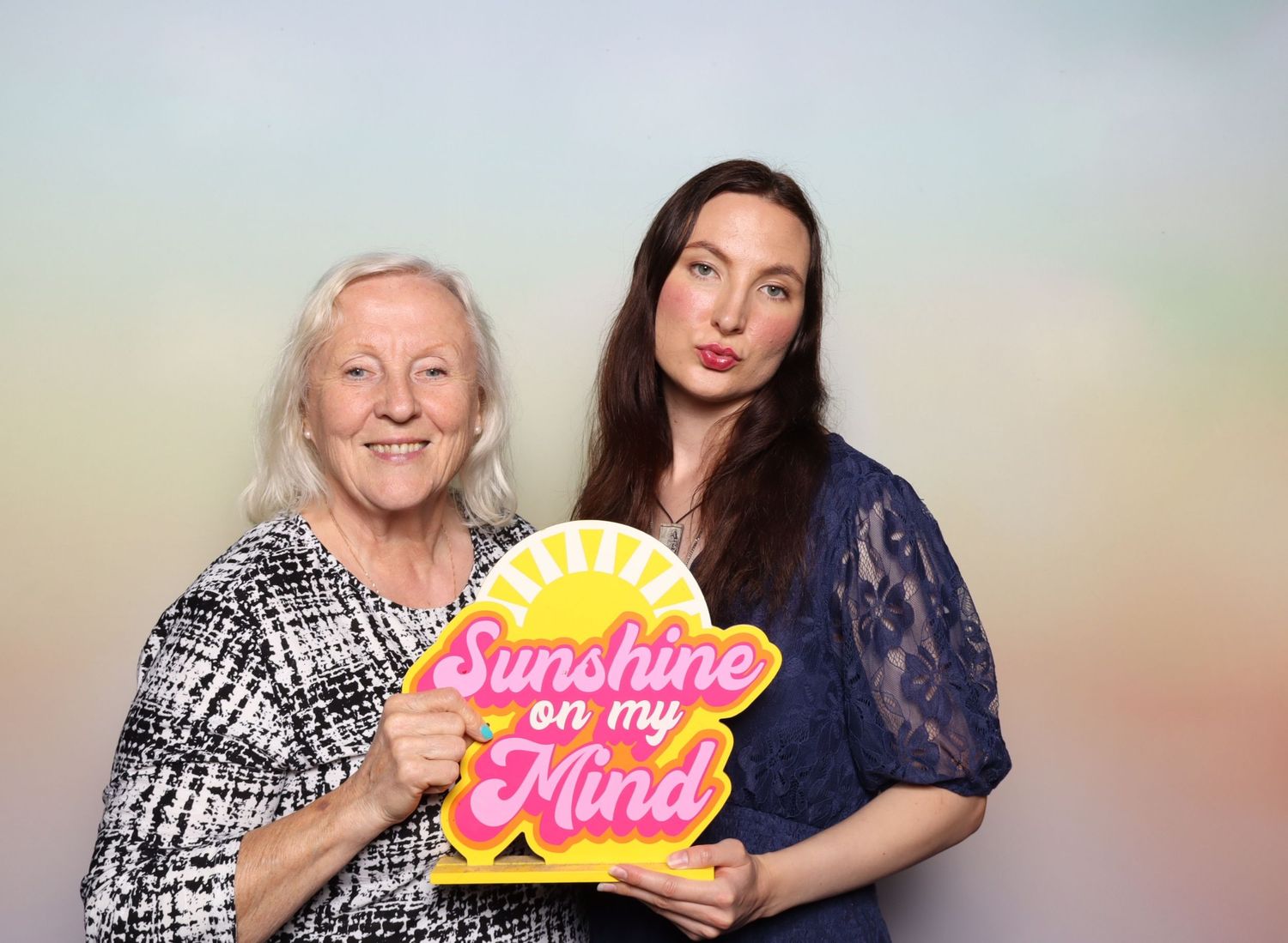 Two women posing together holding a colorful Sunshine on My Mind sign in front of a pastel rainbow background at the Shine in Pleasanton.
