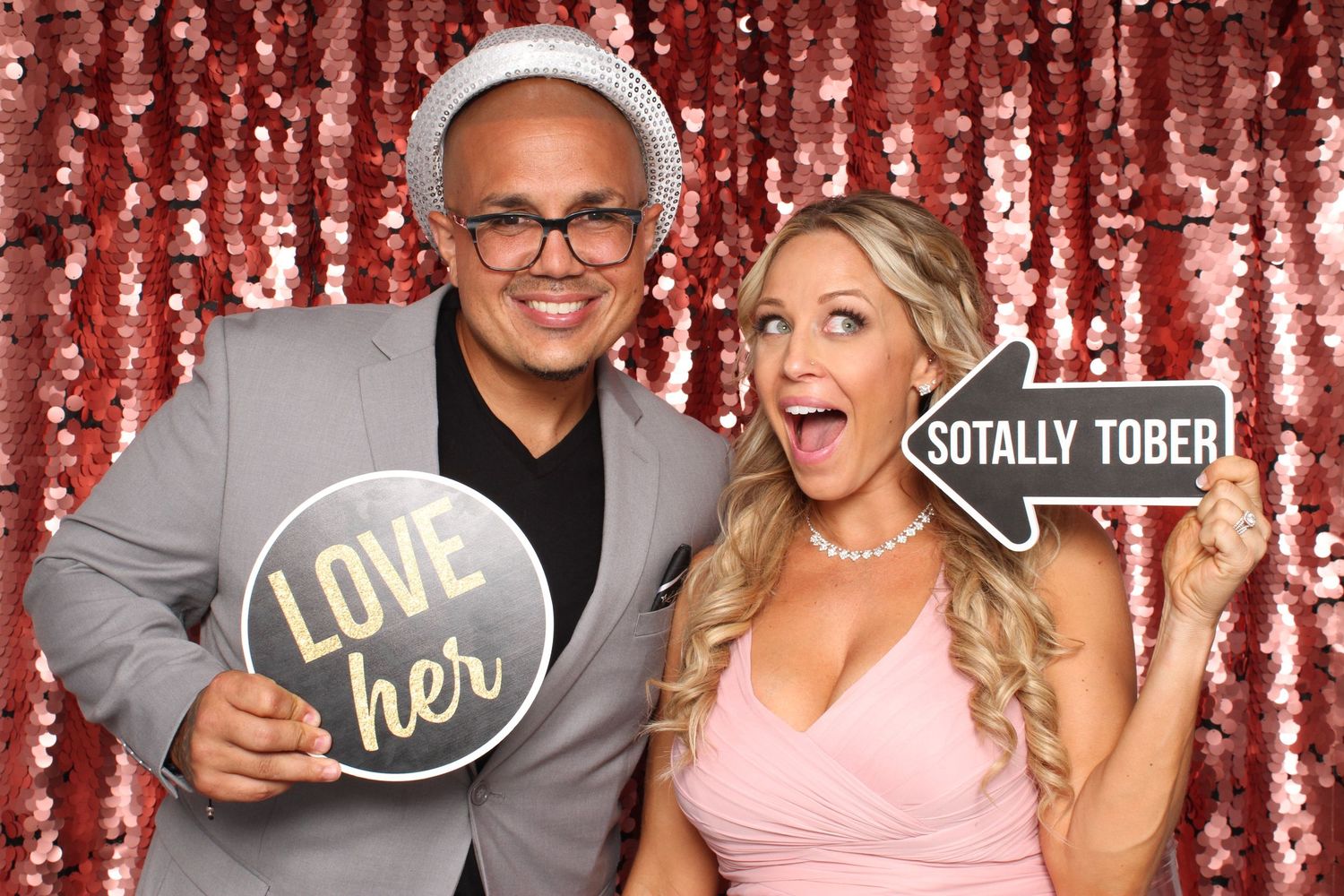 Couple posing at photo booth with fun signs, sequin backdrop, man in gray suit, woman in pink dress.