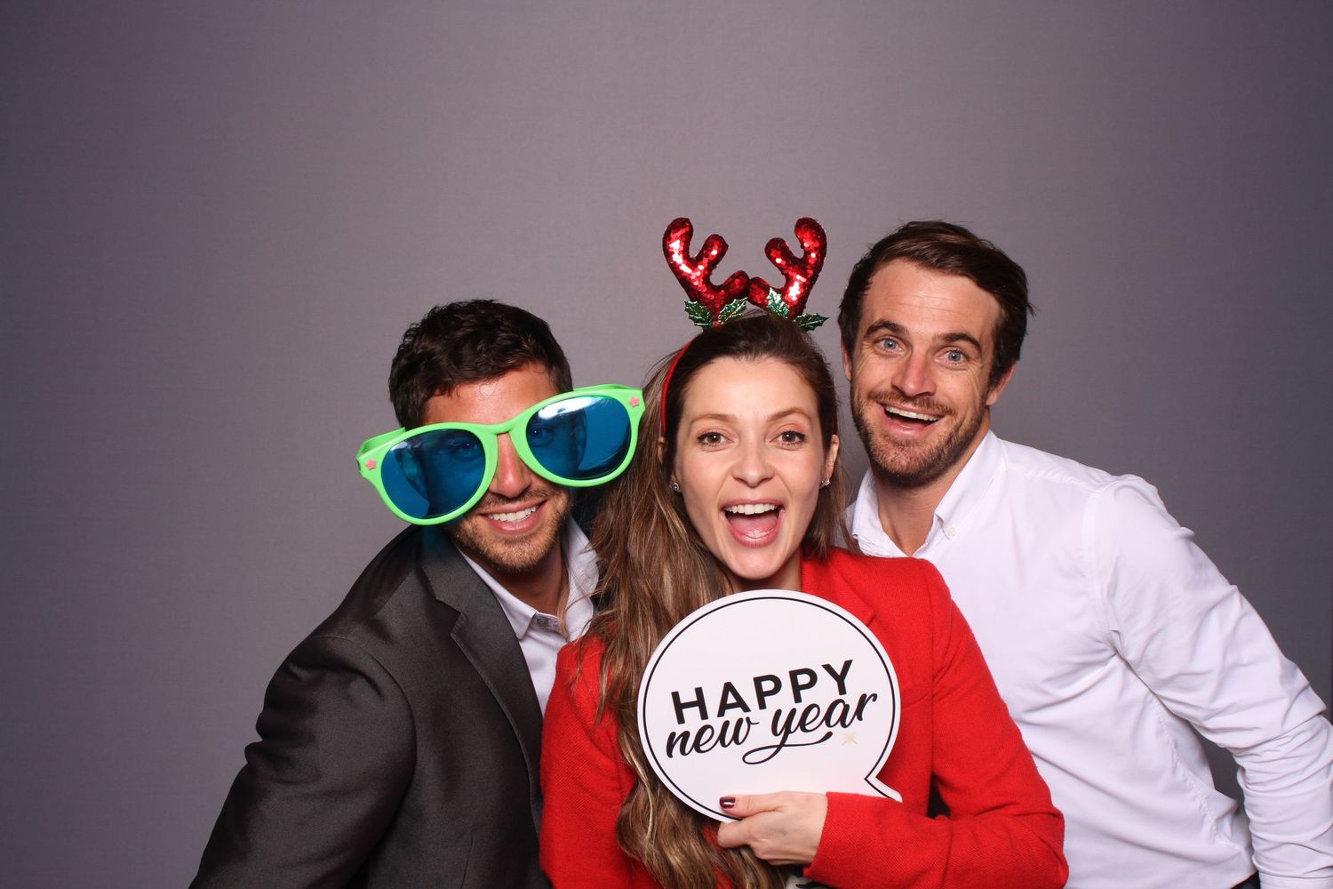 Three friends posing in a photo booth with holiday props, reindeer headband, oversized glasses, and Happy New Year sign.