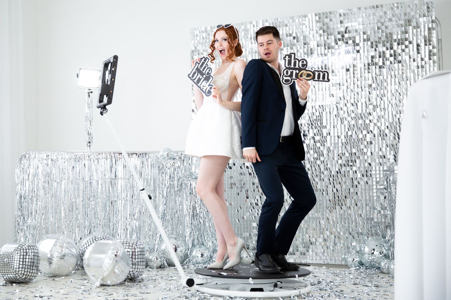 Bride and groom posing at a glittery silver photo booth with props at a wedding reception.