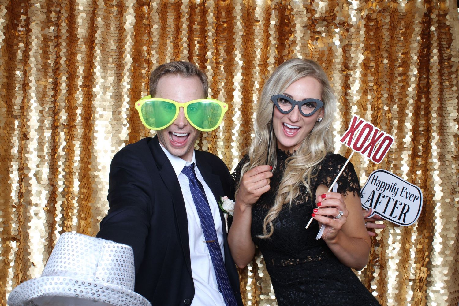 Couple posing in photo booth with oversized glasses and XOXO props against gold sequin backdrop.