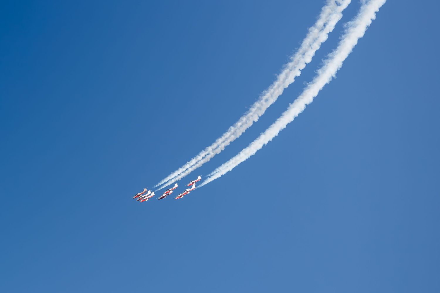 Formation of aircraft performing aerobatic maneuvers with white smoke trails against a clear blue sky.
