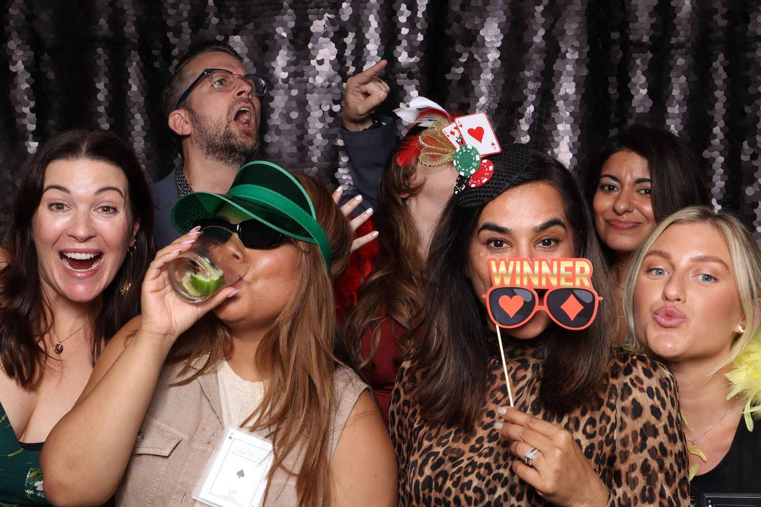 Group of friends posing in a photo booth with fun props including a Winner sign and heart glasses.