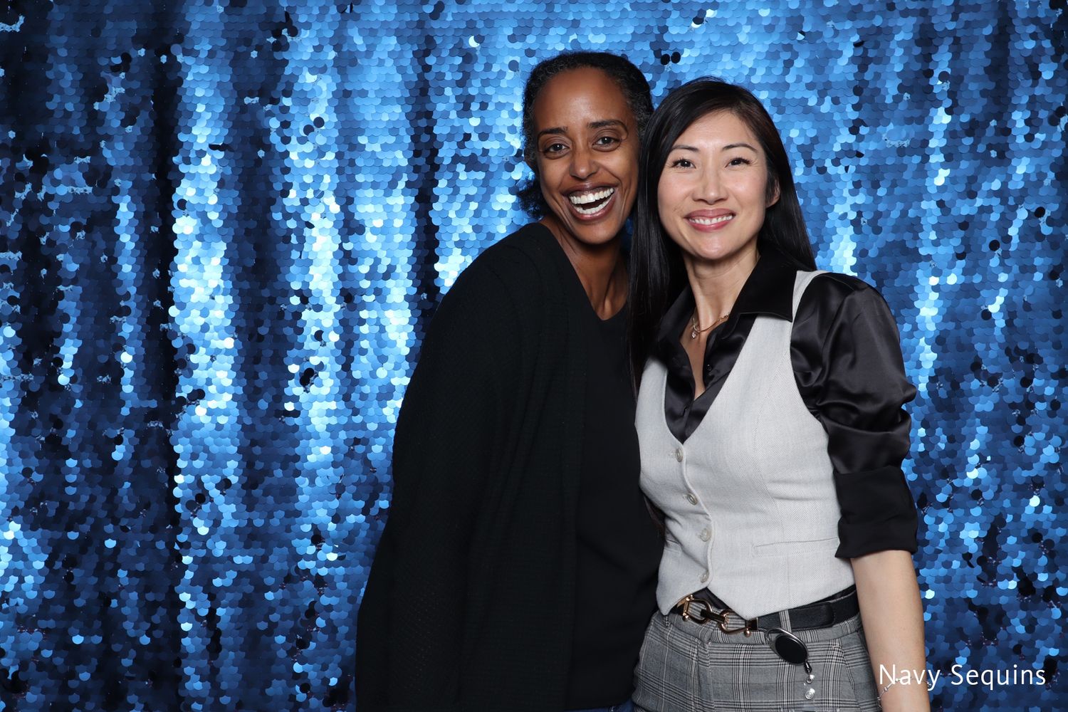 Two women smiling together in front of blue sequin photo booth backdrop.
