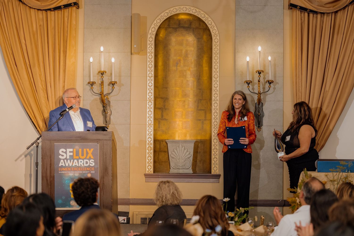 Woman accepts award at SF UX Awards ceremony on elegant stage with candles and gold decor.