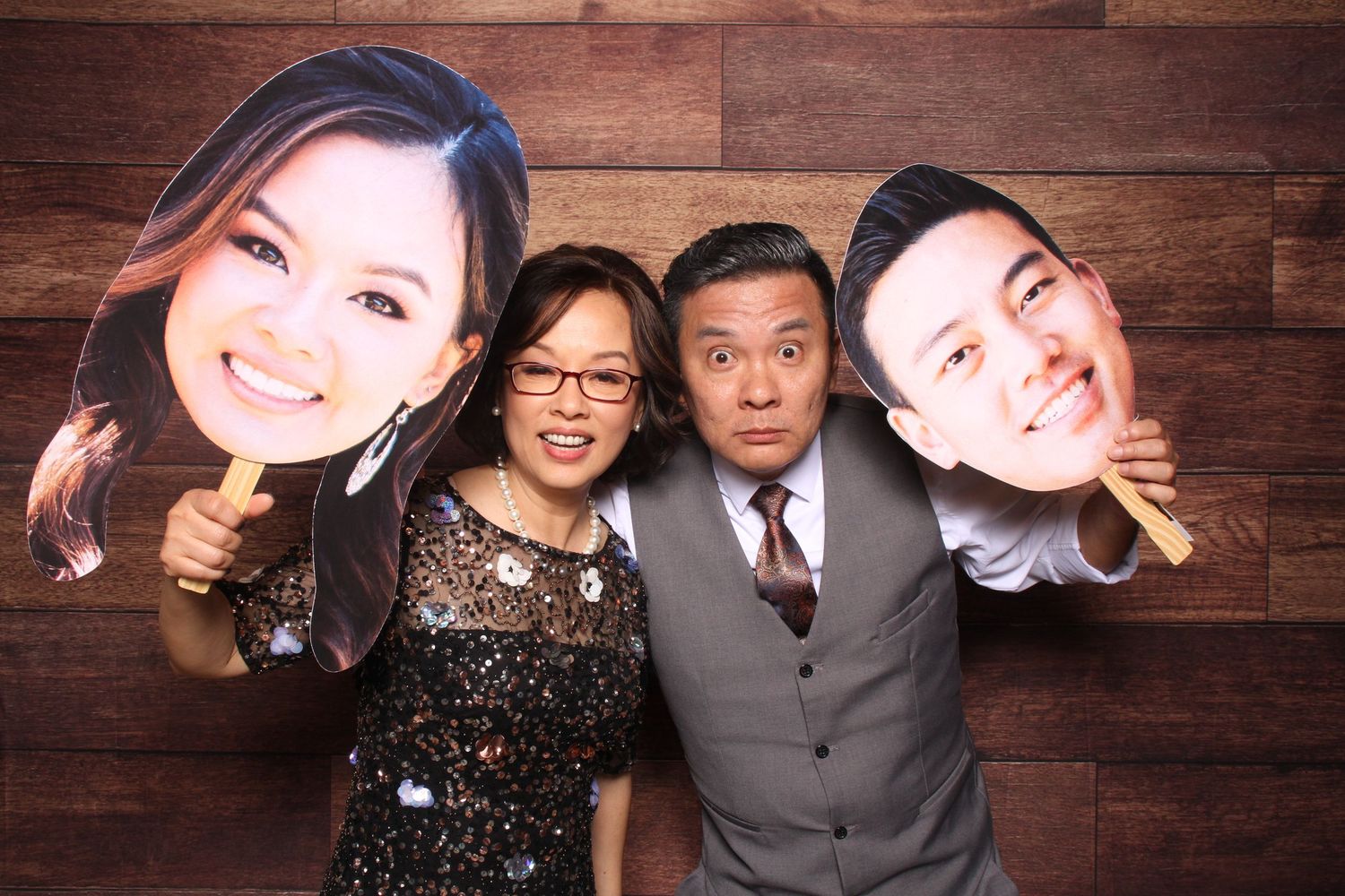 Two people posing at a photo booth holding large cutout face props on wooden background in a party in Oakland.