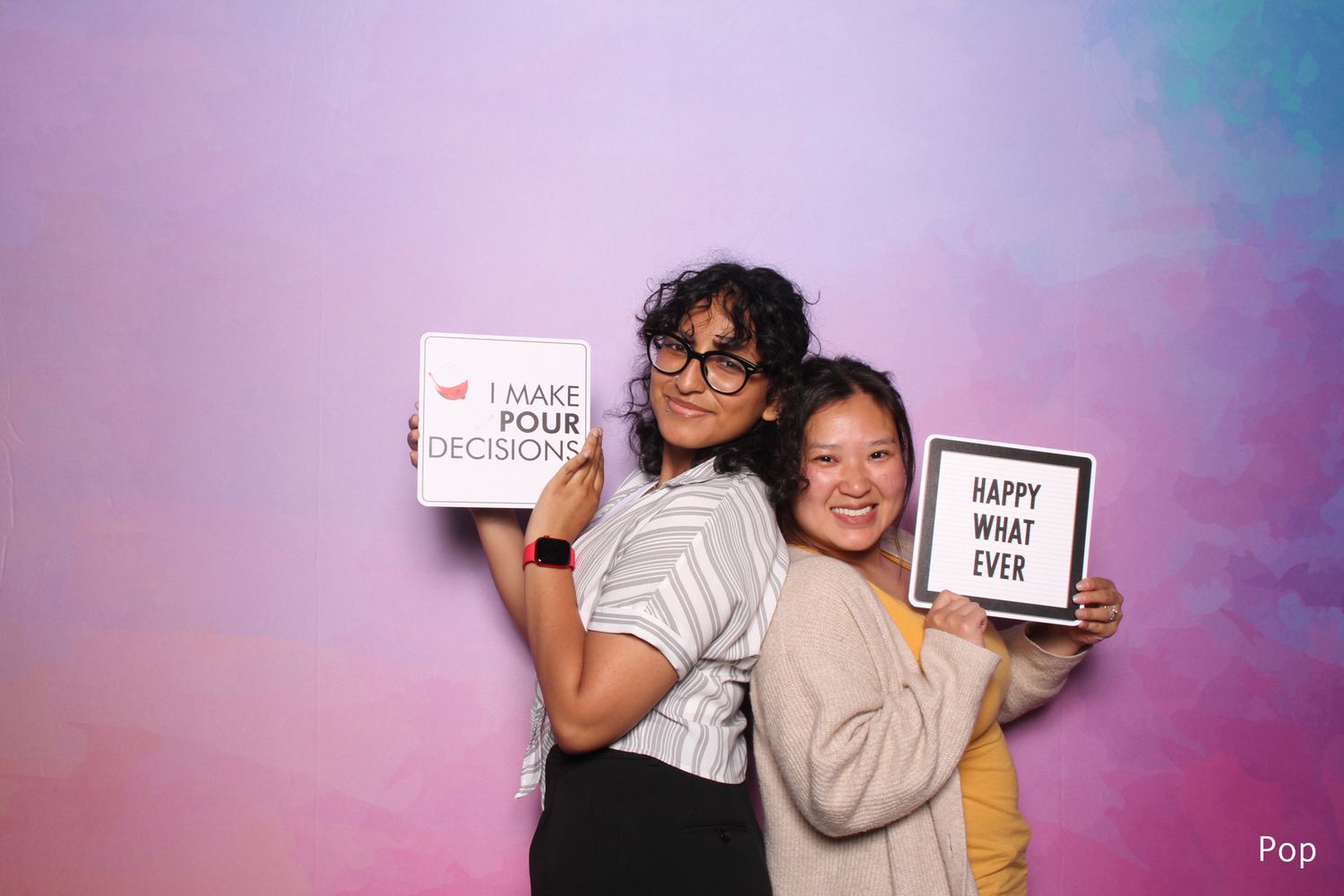 Two women holding funny signs at colorful pastel photo booth backdrop.