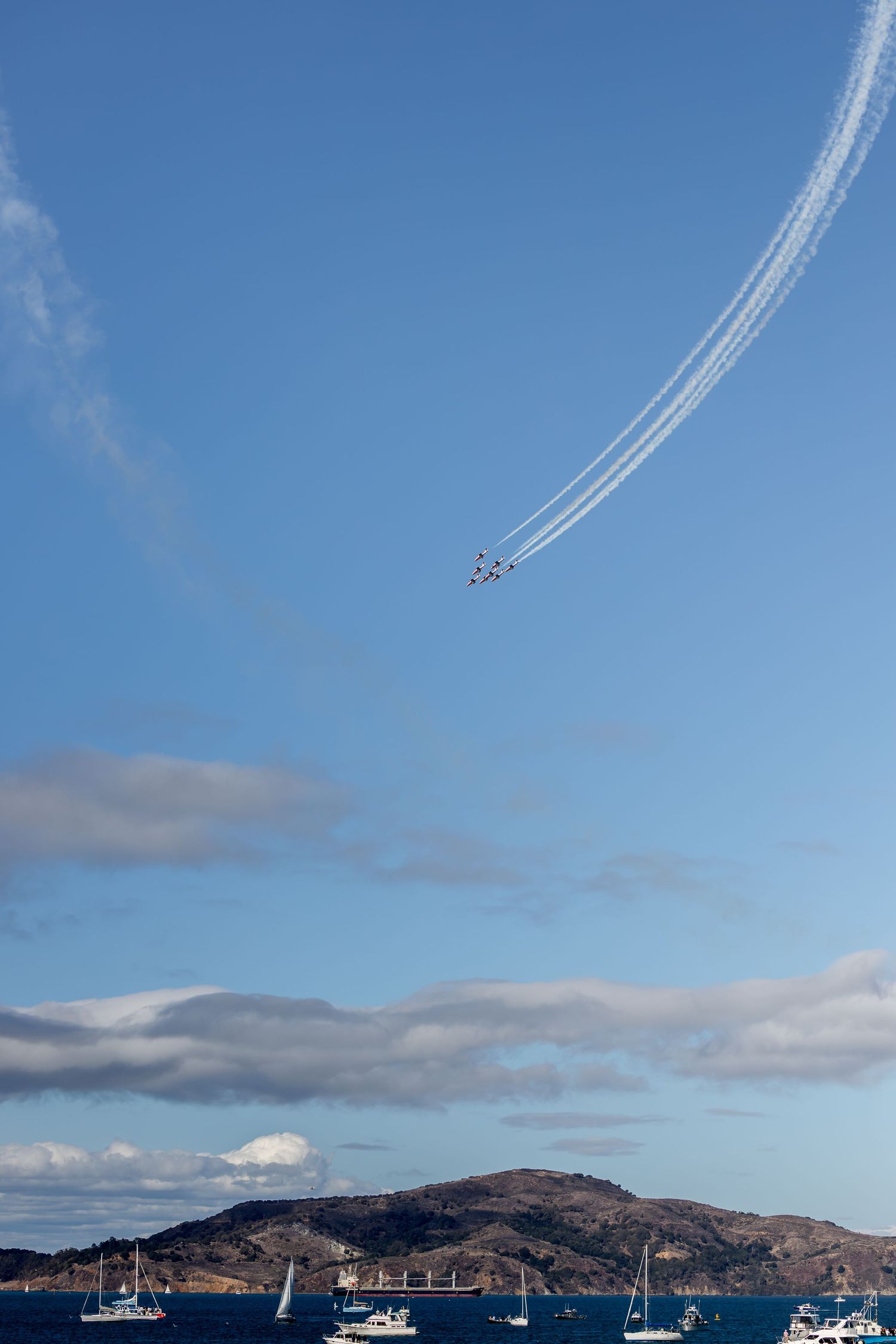 Formation of jets performing aerobatic maneuvers over a harbor with sailboats and hills under a blue sky.