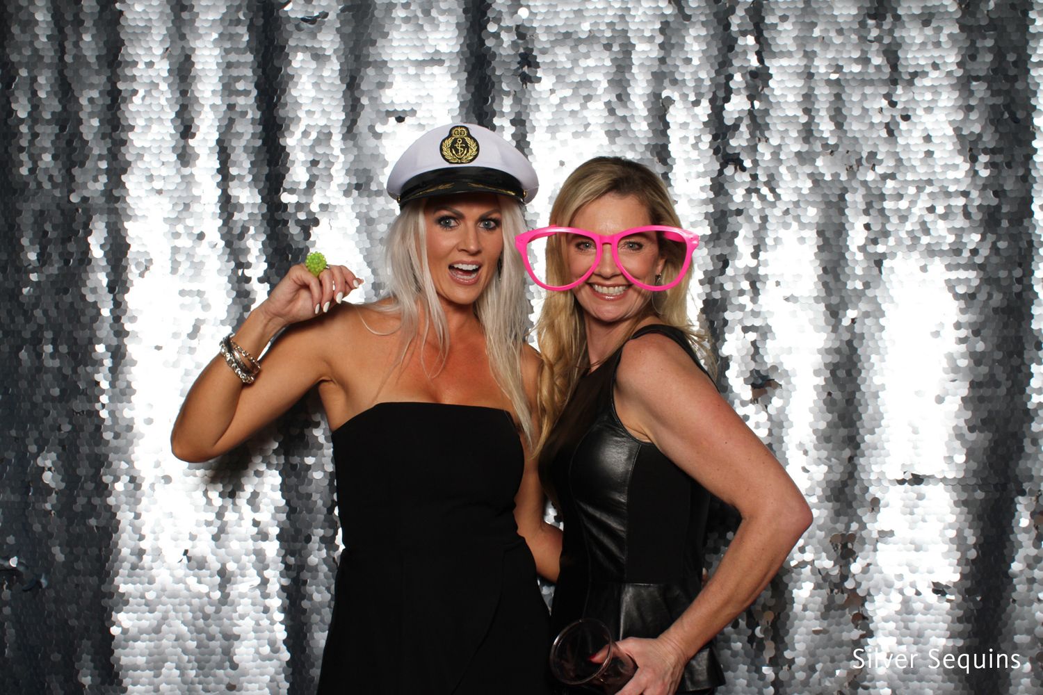 Two women posing in a photo booth with silver sequin backdrop, one wearing a captain hat, the other pink oversized glasses.