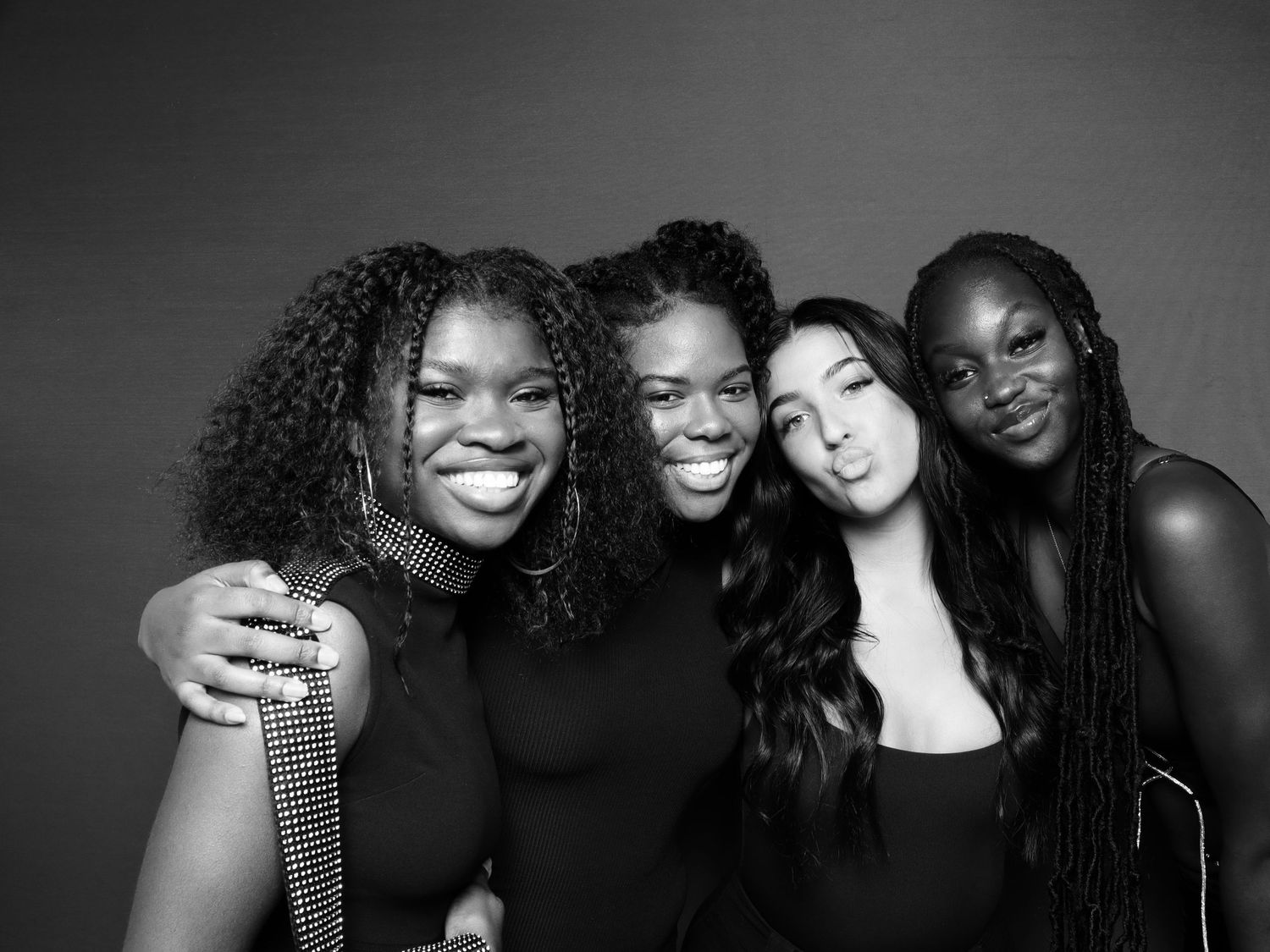 Four young women smiling together in a black and white portrait photo, wearing stylish outfits.