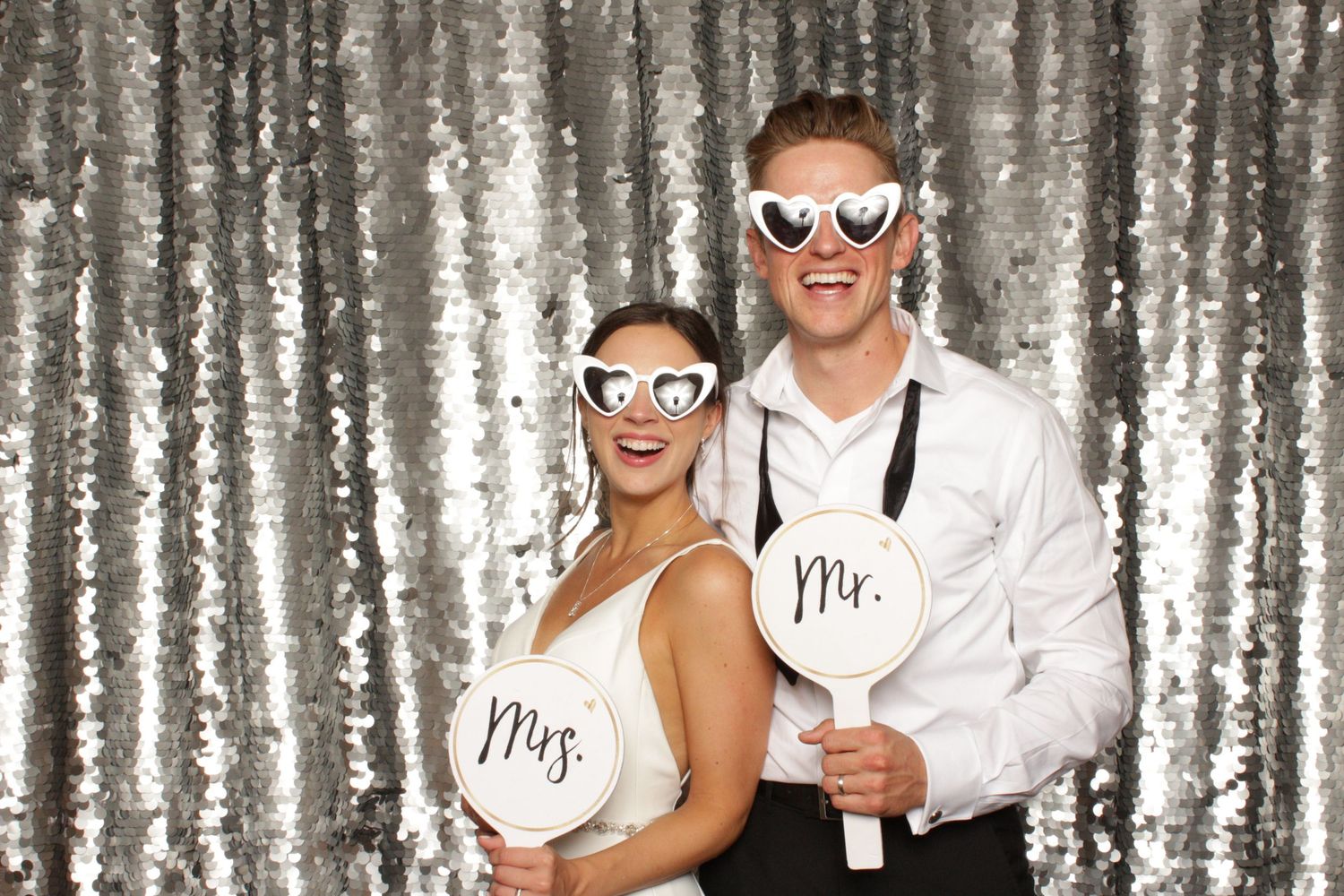 Newlyweds in a wedding photo booth wearing heart-shaped glasses against a silver sequin backdrop.