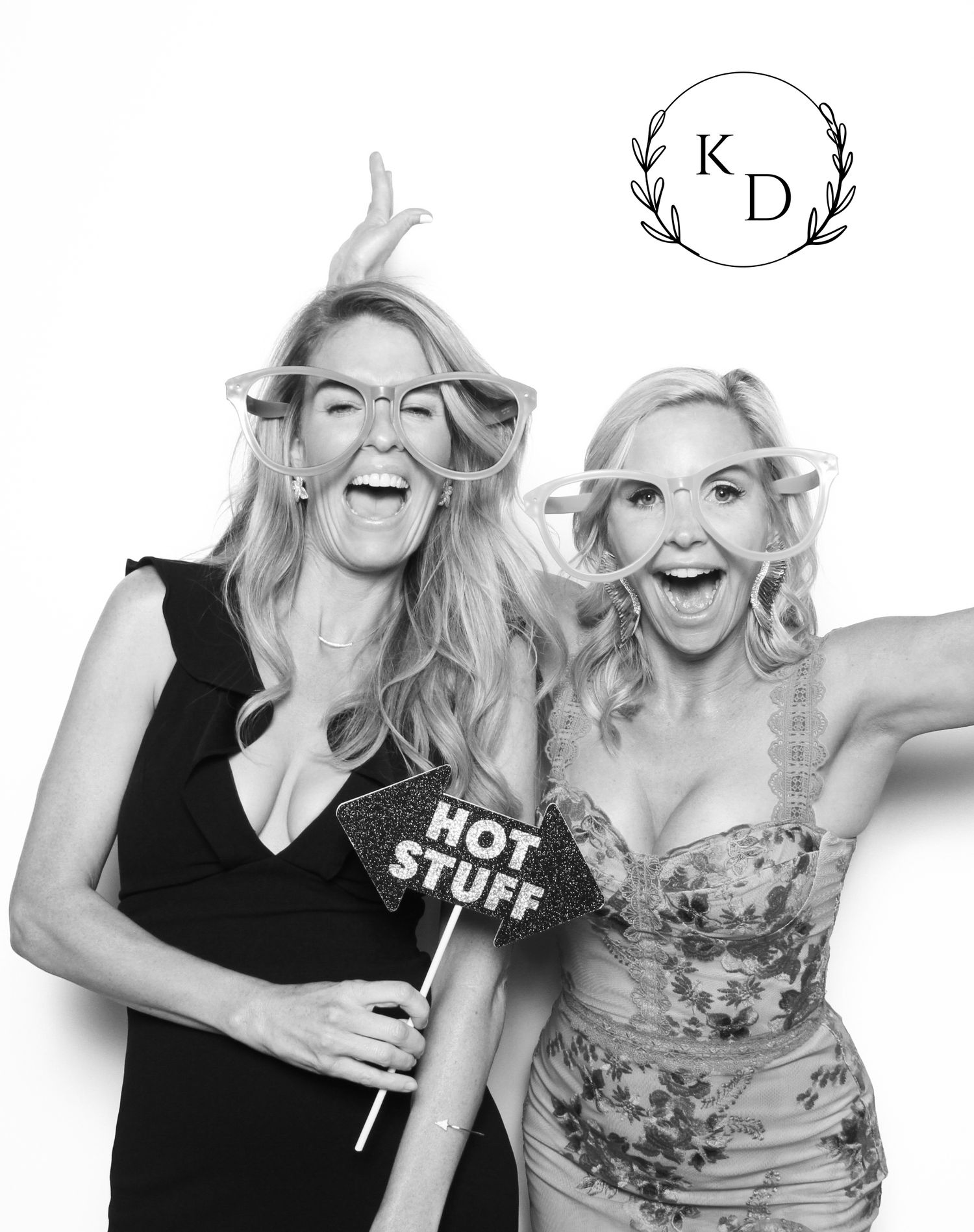 Two blonde women laughing and posing in a photo booth with fun props, black and white wedding photo.