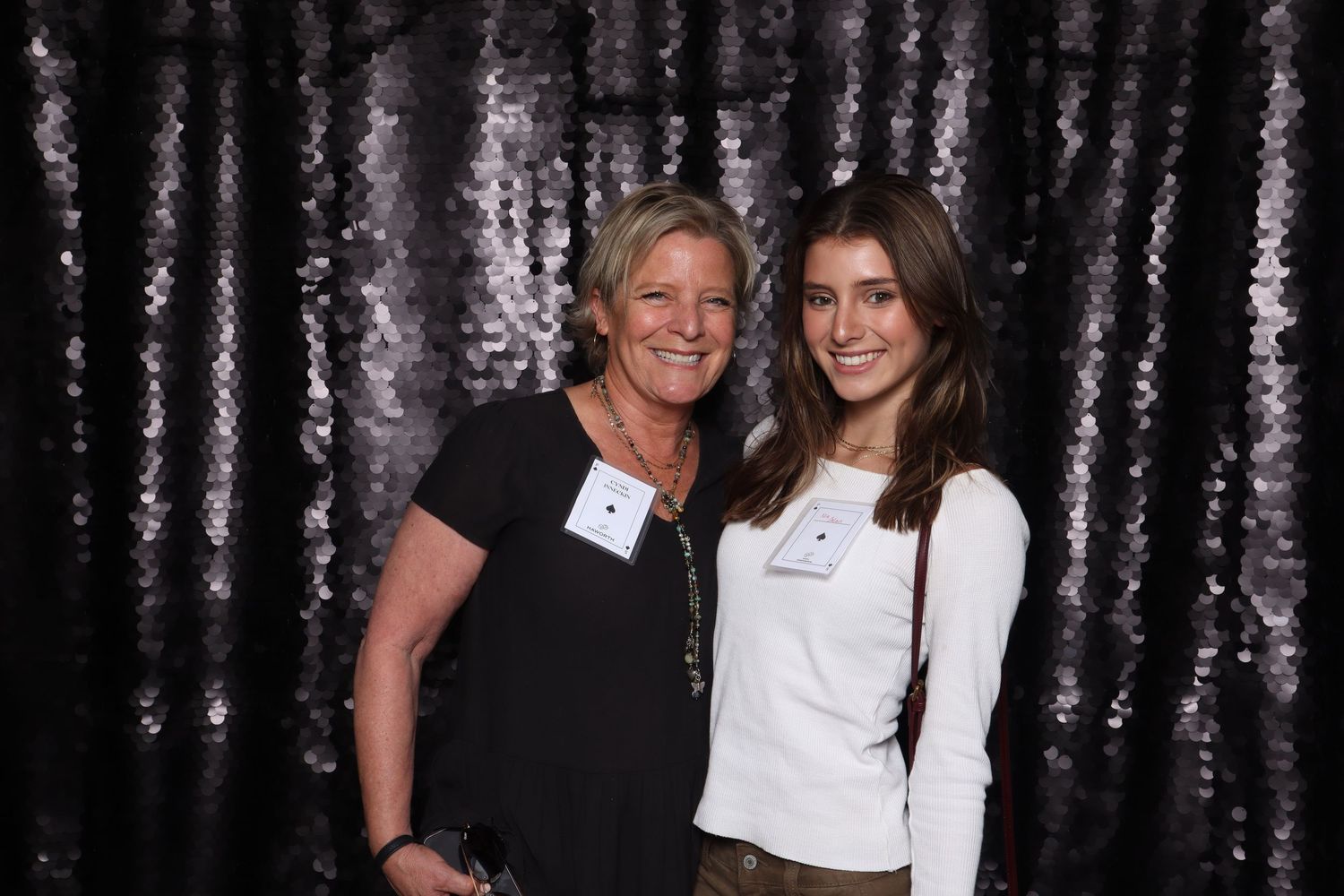 Two women smiling together in front of a black sequin backdrop at a formal event, wearing name badges.