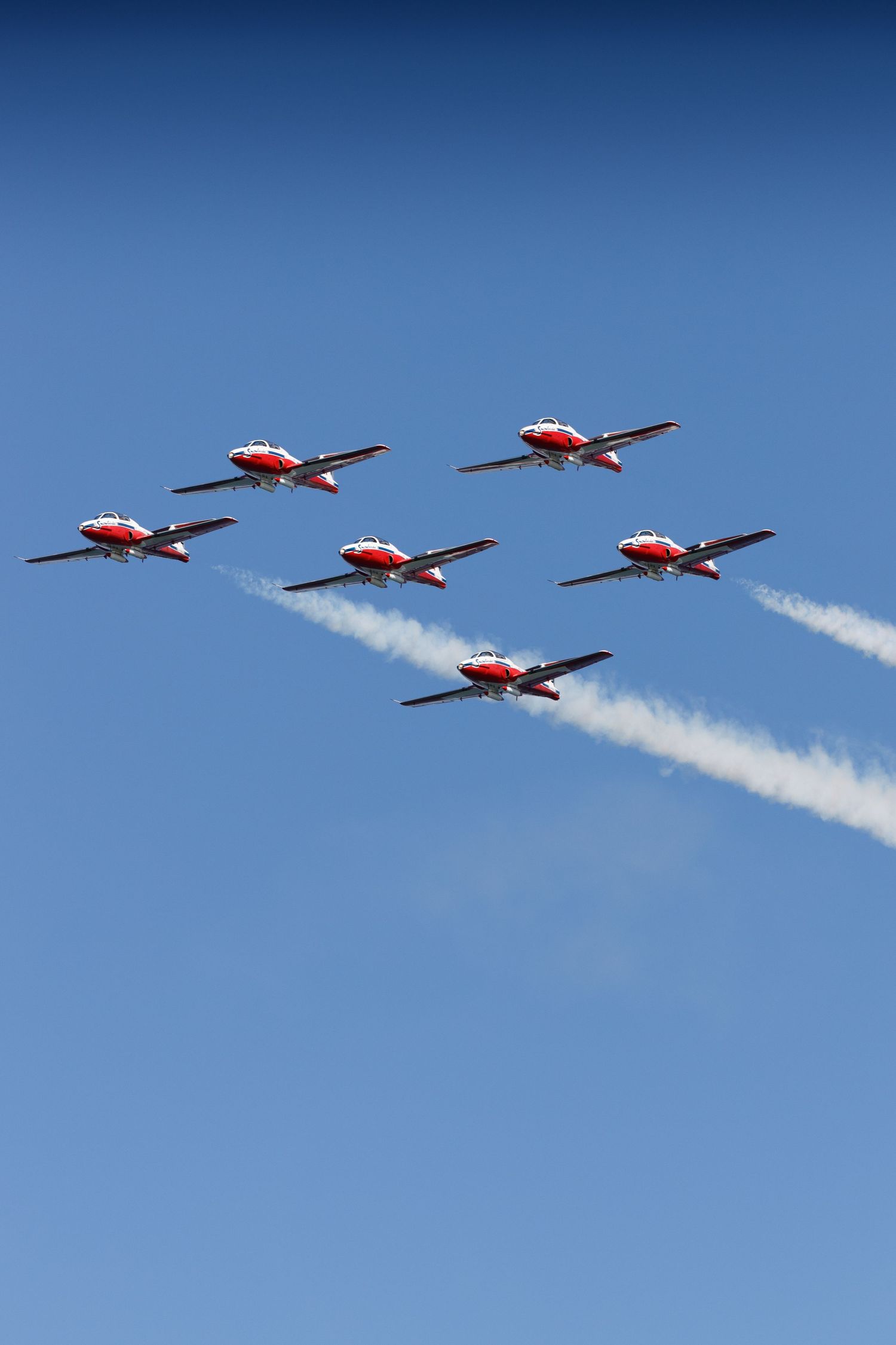 Six red and white aerobatic jets fly in tight diamond formation against a clear blue sky, trailing white smoke.