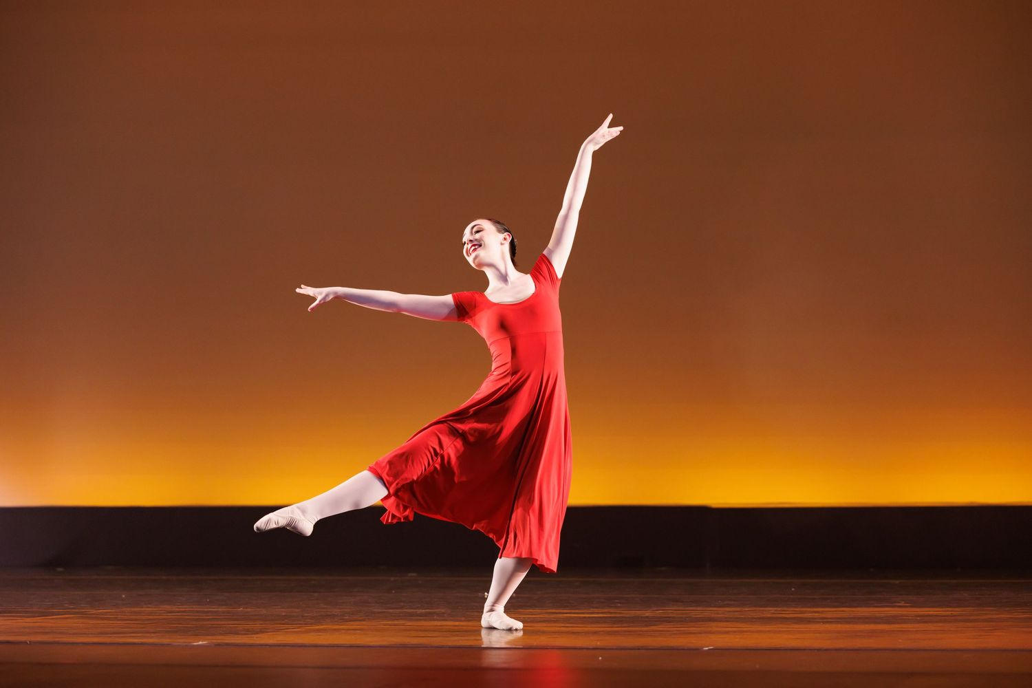 A graceful ballet dancer in a red dress performs an elegant arabesque pose on a warmly lit stage.  Las Positas College