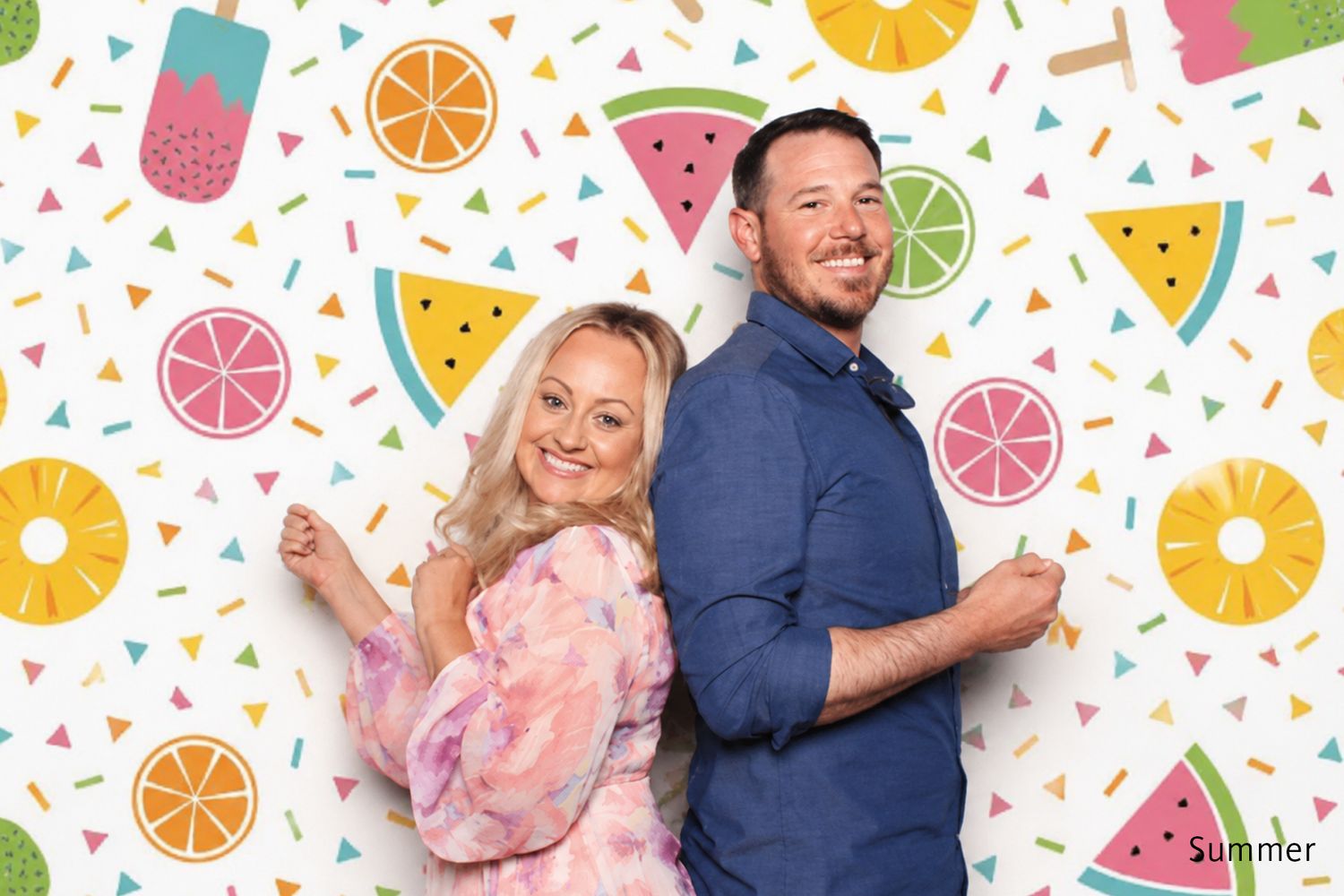 Couple posing and smiling in front of a colorful summer fruit photo booth backdrop.