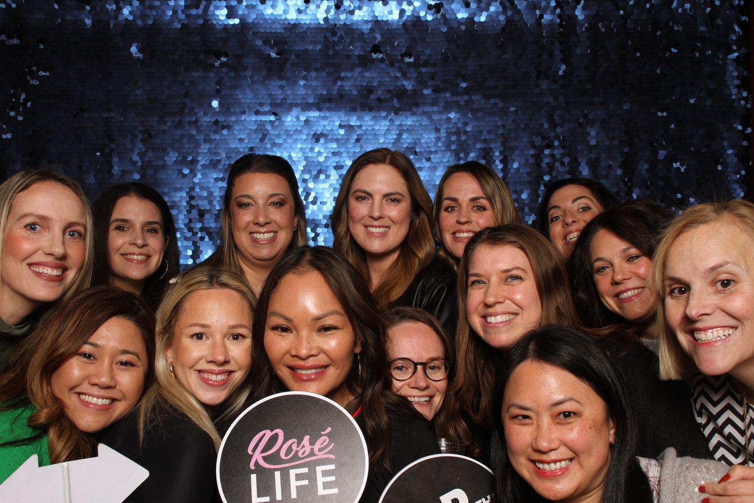 Group of smiling women posing together at a party, one holding a Rose Life sign, with a glittery backdrop.