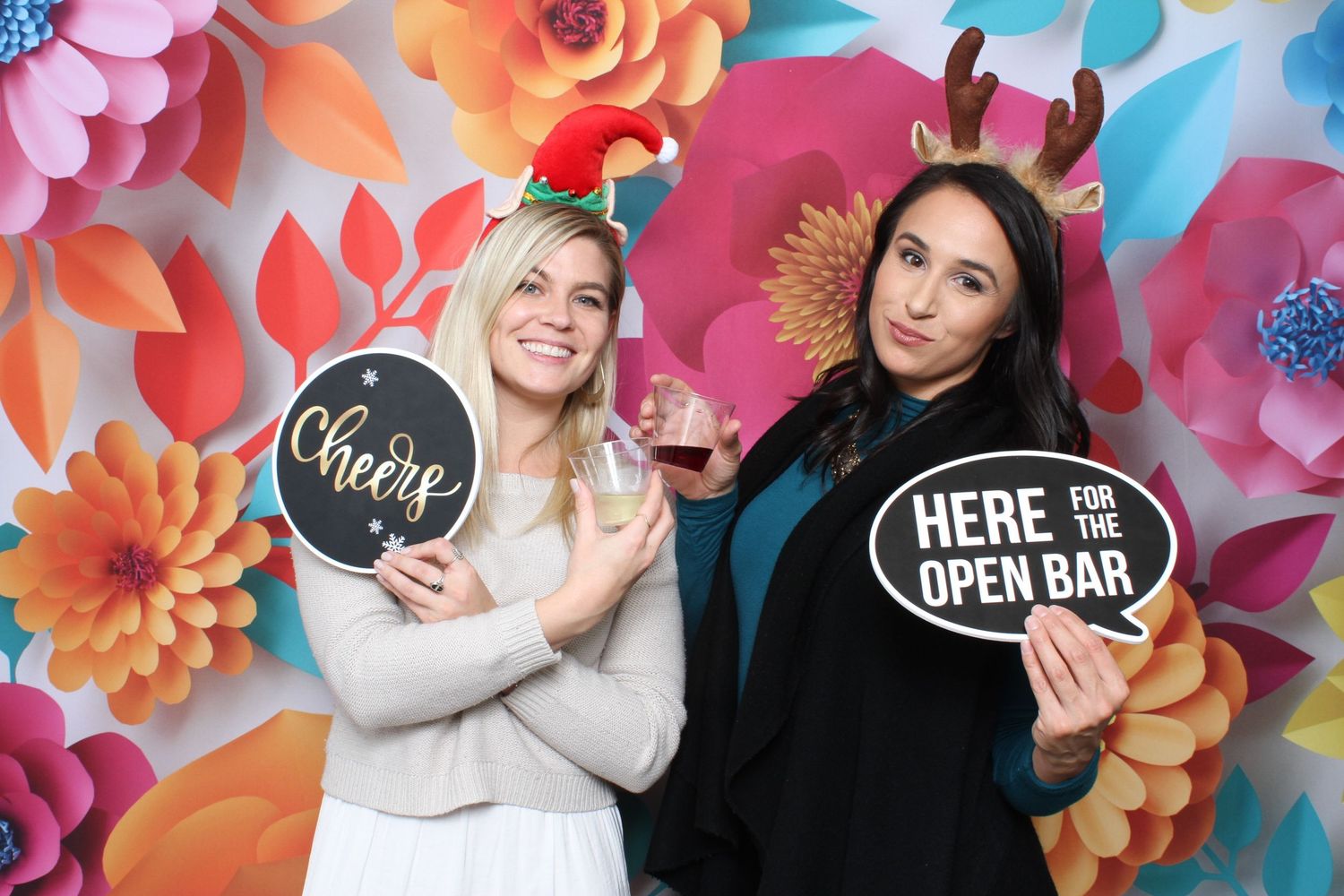 Two women pose at a festive photo booth with holiday headbands, holding fun signs reading Cheers and Here for the Open Bar.