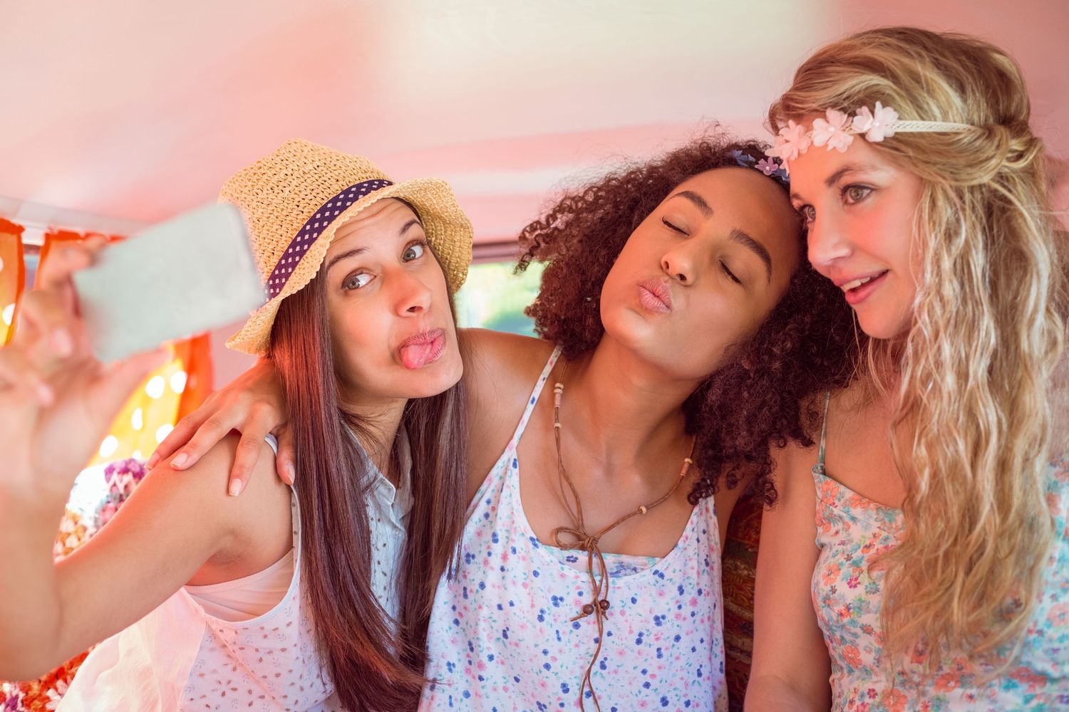 Three young women making duck faces while taking a selfie inside a colorful van at a summer festival.