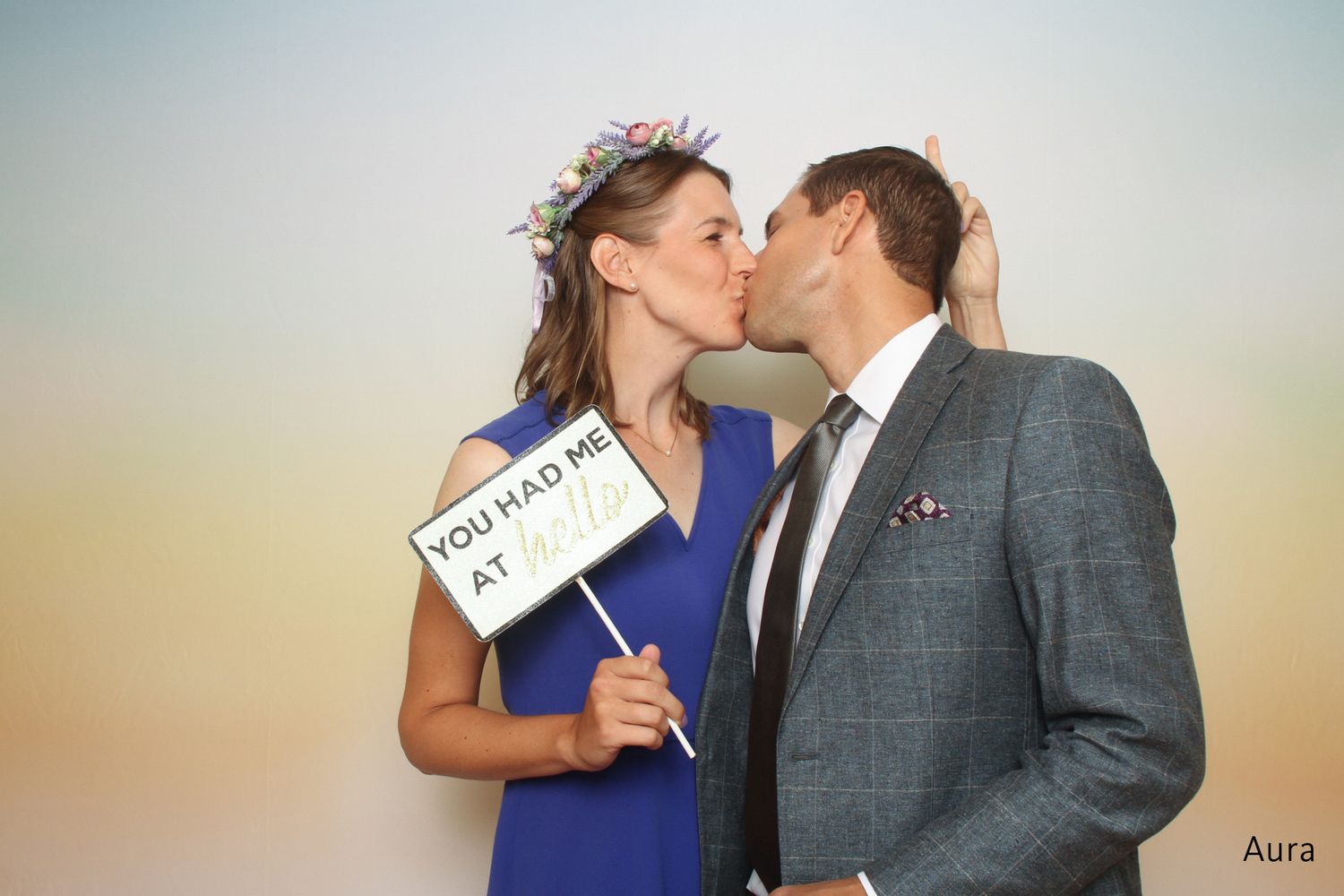 Couple kissing while holding a You Had Me At Hello sign in front of an Aura backdrop.