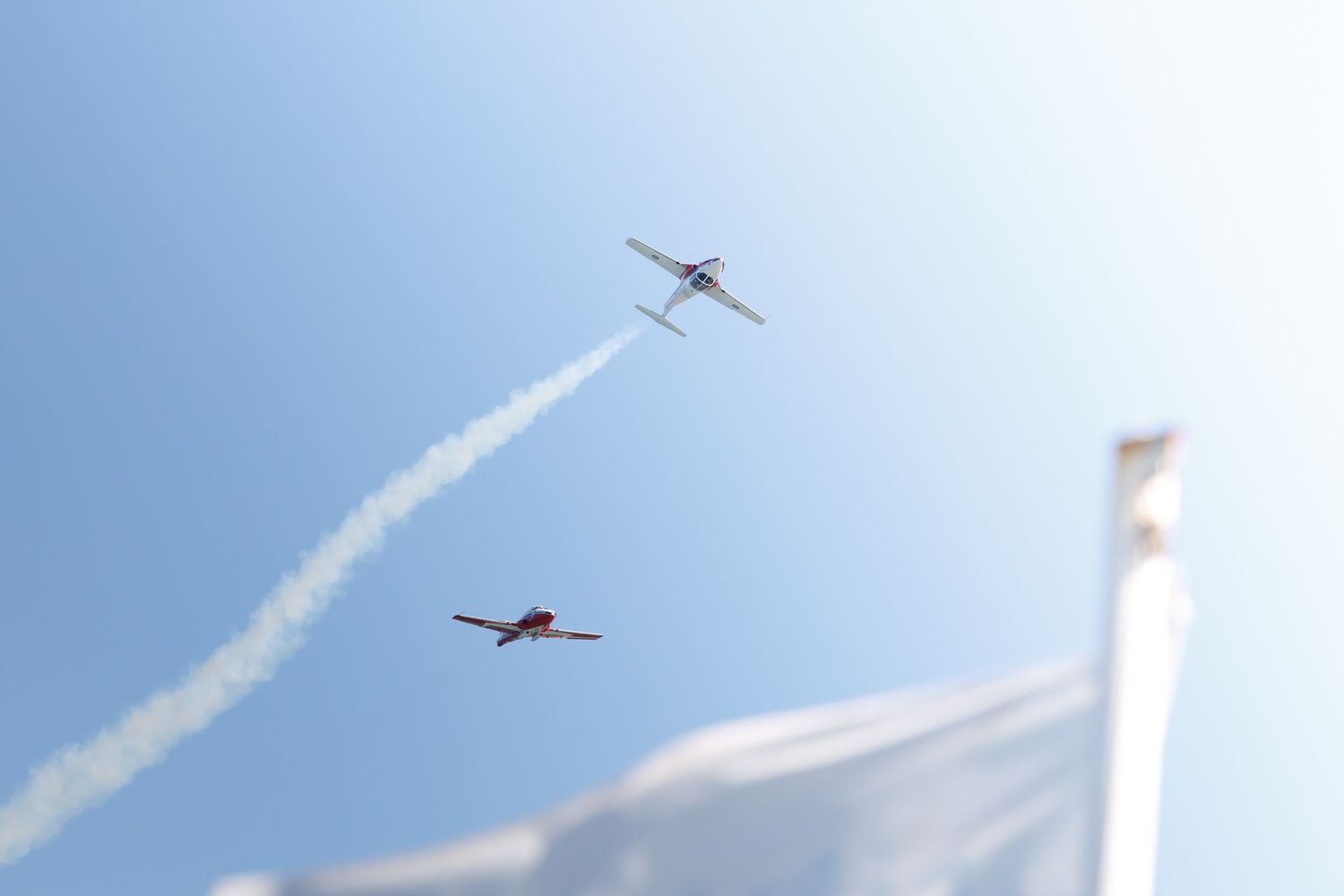 Two aircraft performing aerobatic maneuvers leaving white smoke trails against a clear blue sky at an airshow.