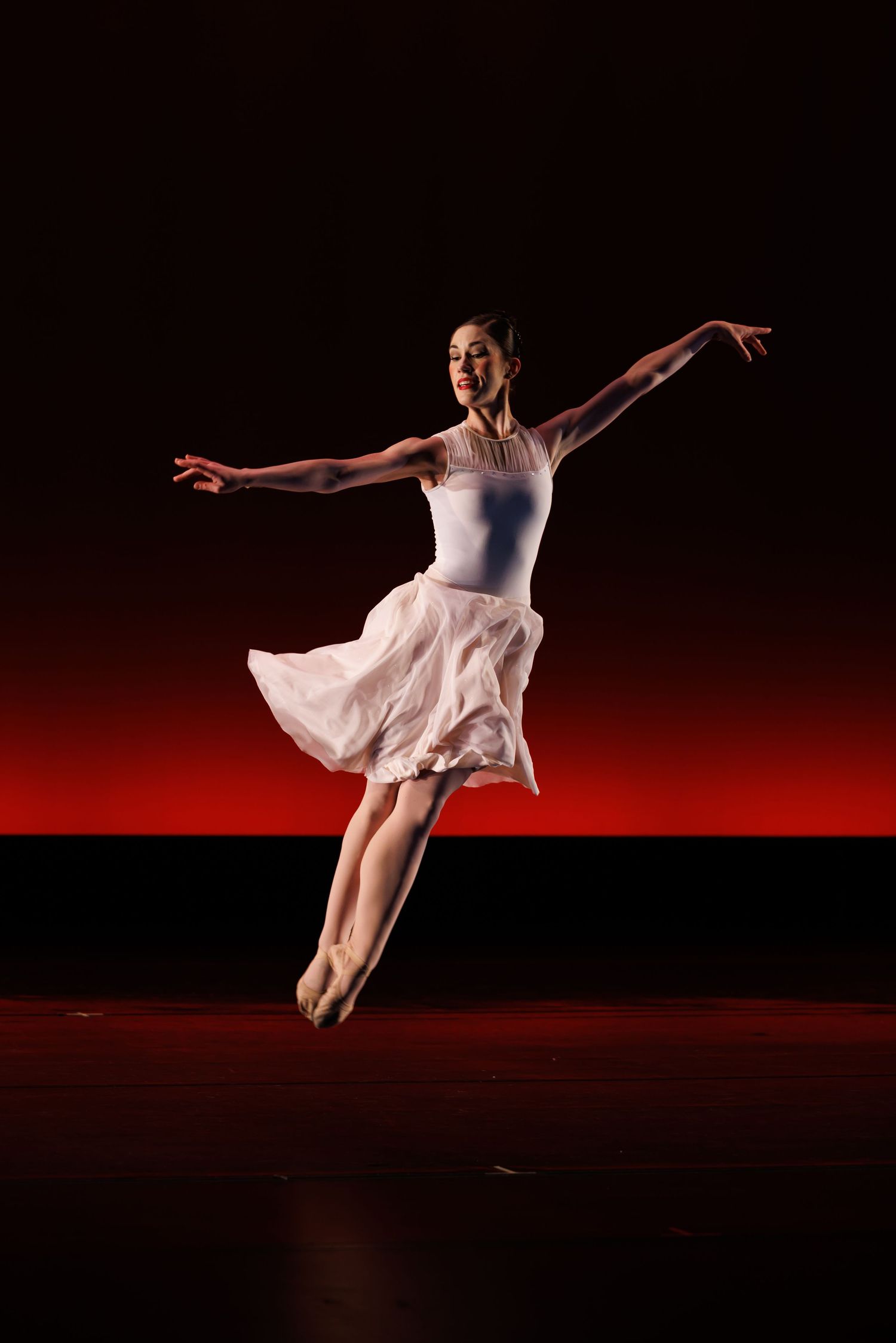 A graceful ballerina in a white tutu performs an elegant arabesque on a dark stage with a dramatic red backdrop.  Las Positas College