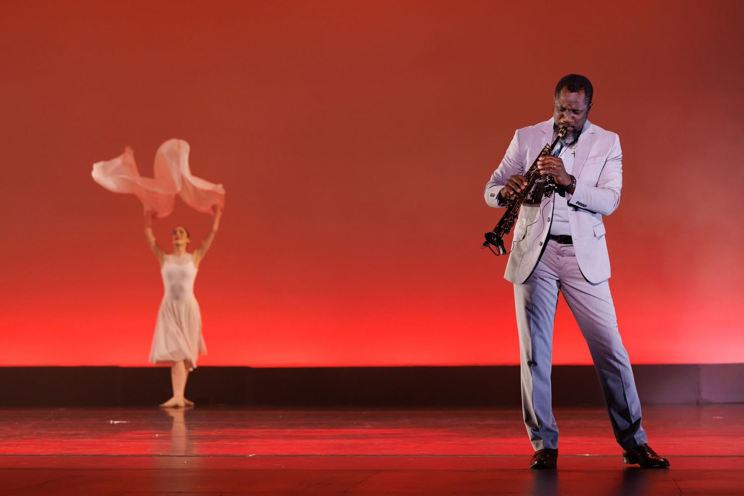 A saxophonist in a white suit performs on a red-lit stage while a dancer in white moves gracefully behind him. Las Positas