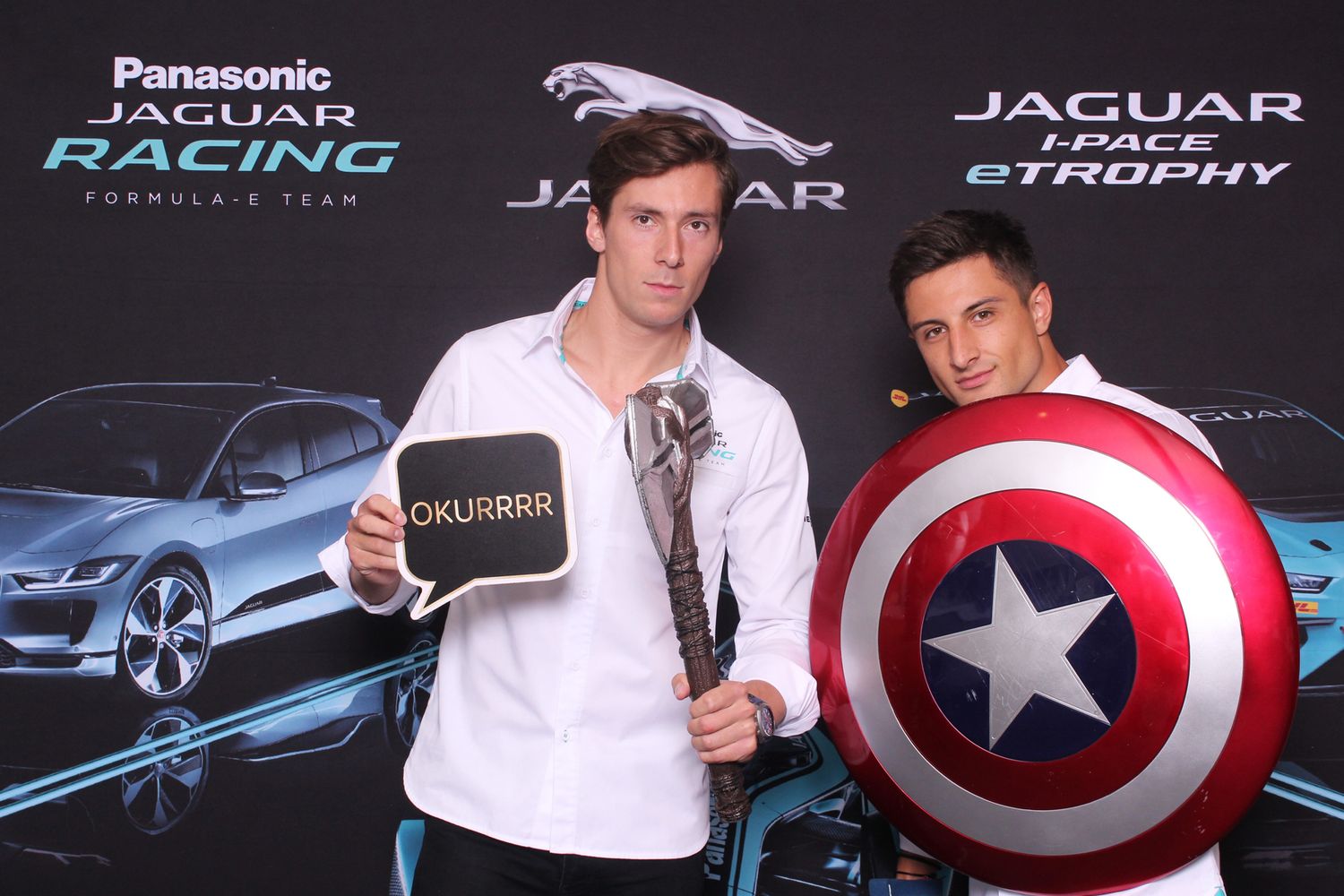 Two men pose at Jaguar Racing Formula E event, one holds Captain America shield, other holds OKURRR sign.