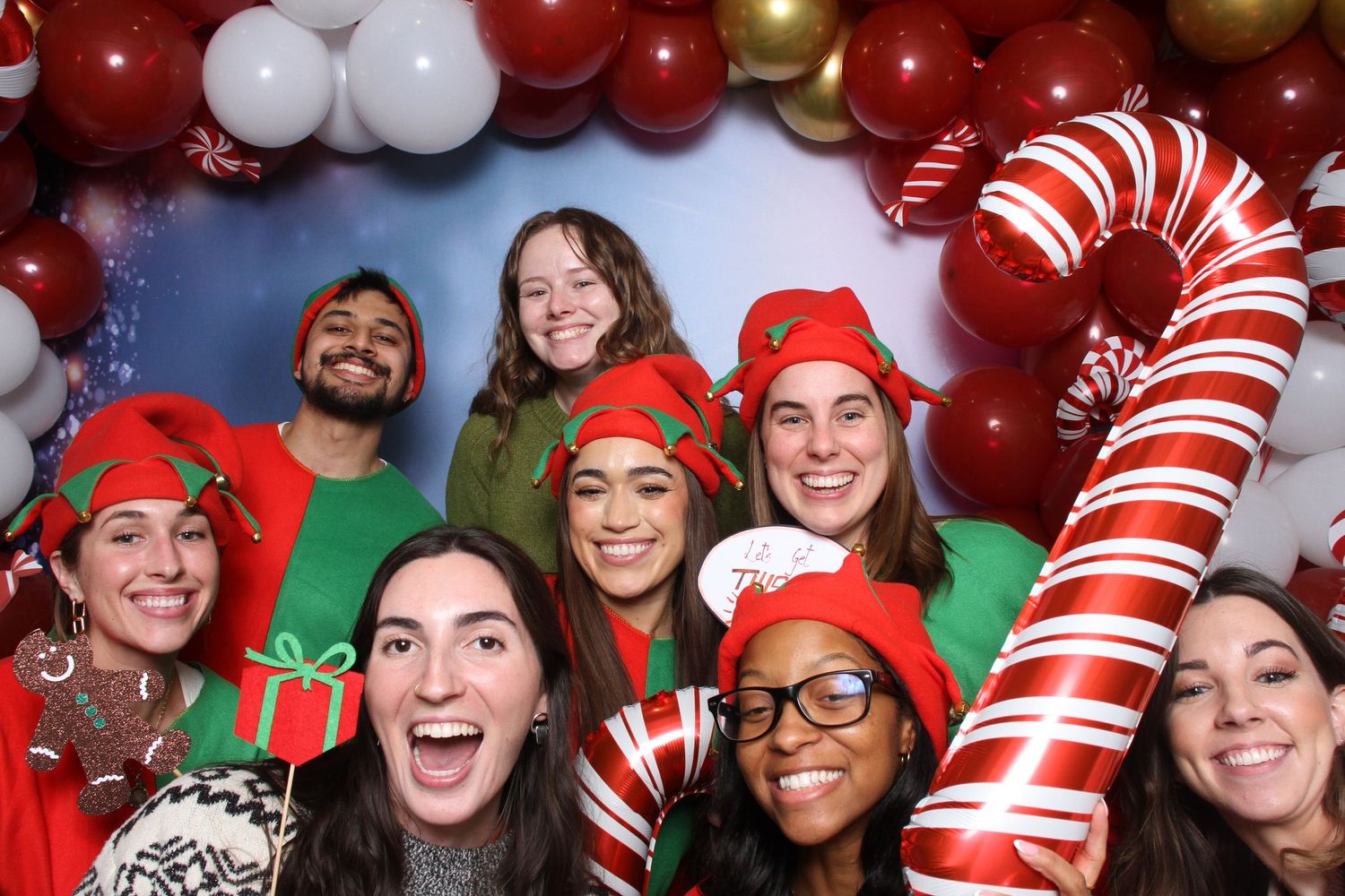 Group of friends posing at a Christmas photo booth with elf hats, candy cane props, and festive balloon decorations.