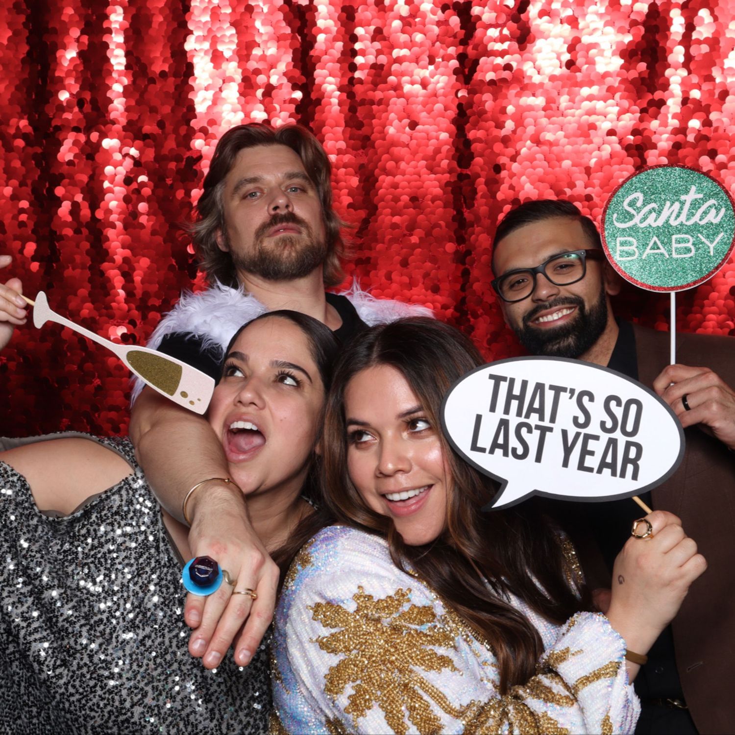 Four friends posing at a festive photo booth with red sequin backdrop, holding fun holiday signs at a Christmas party. Gensler Holiday Party