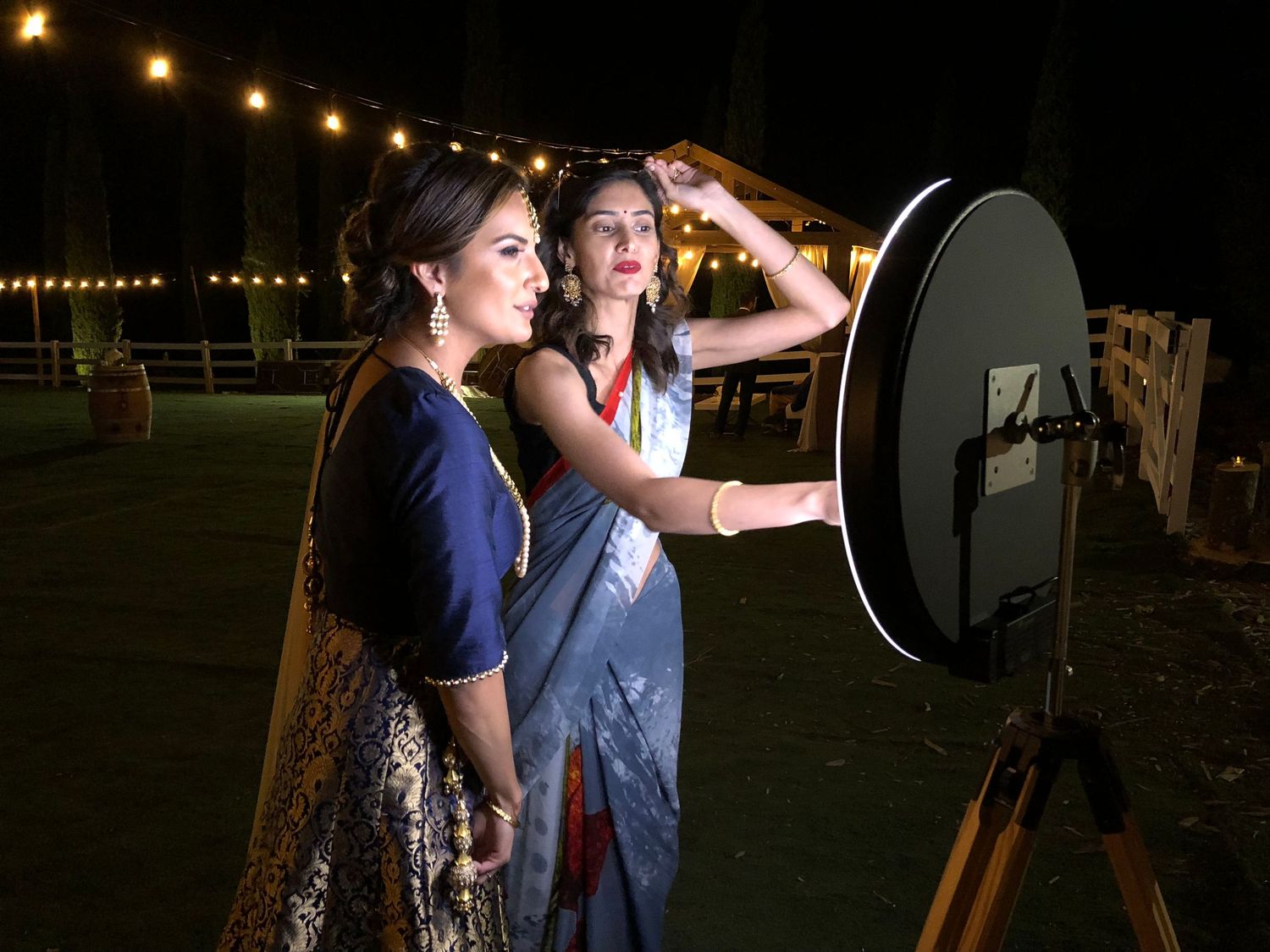 Two women in elegant sarees pose near a circular ring light on a tripod at an outdoor evening event.