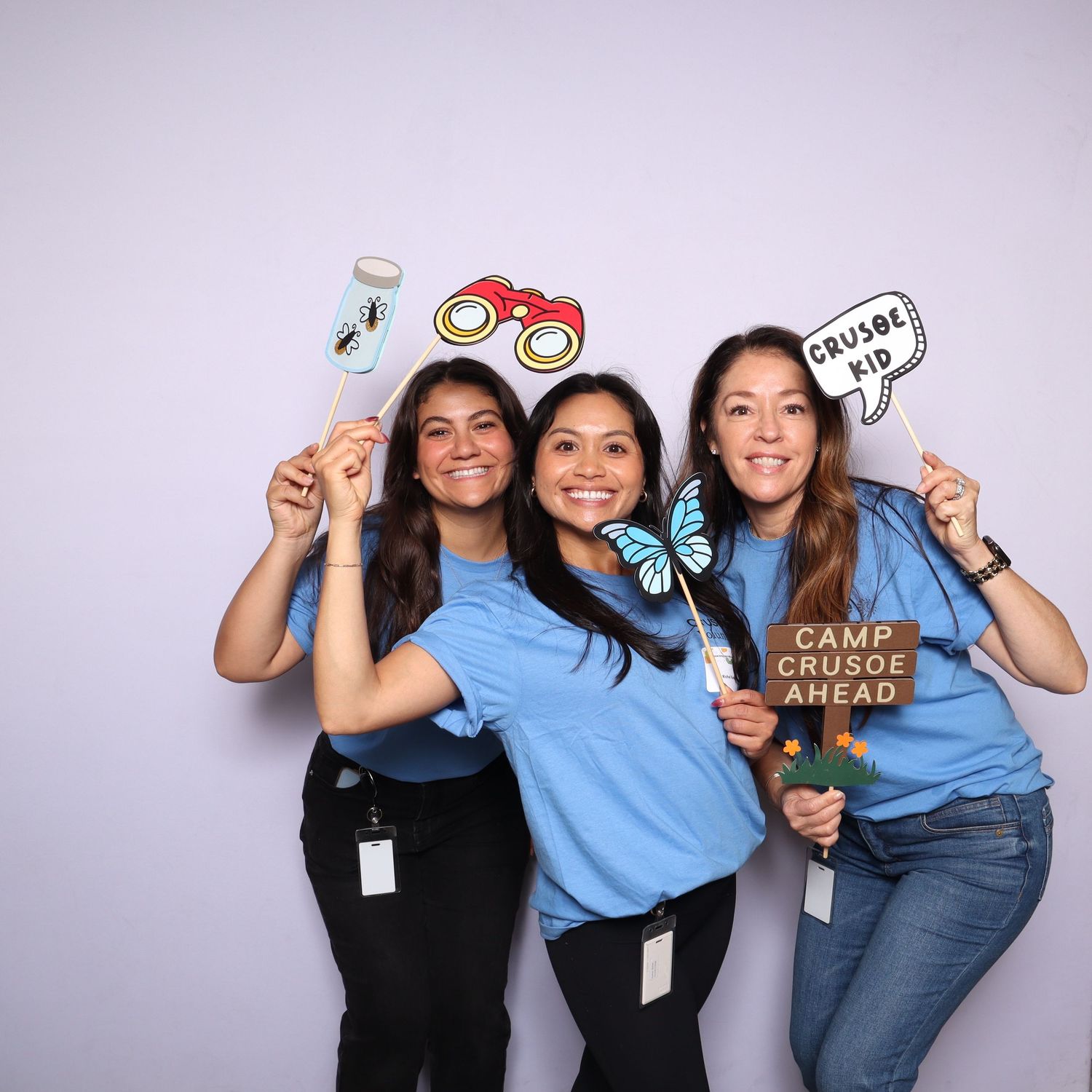 Three women in blue shirts smile holding Camp Crusoe photo booth props including binoculars and butterfly signs.