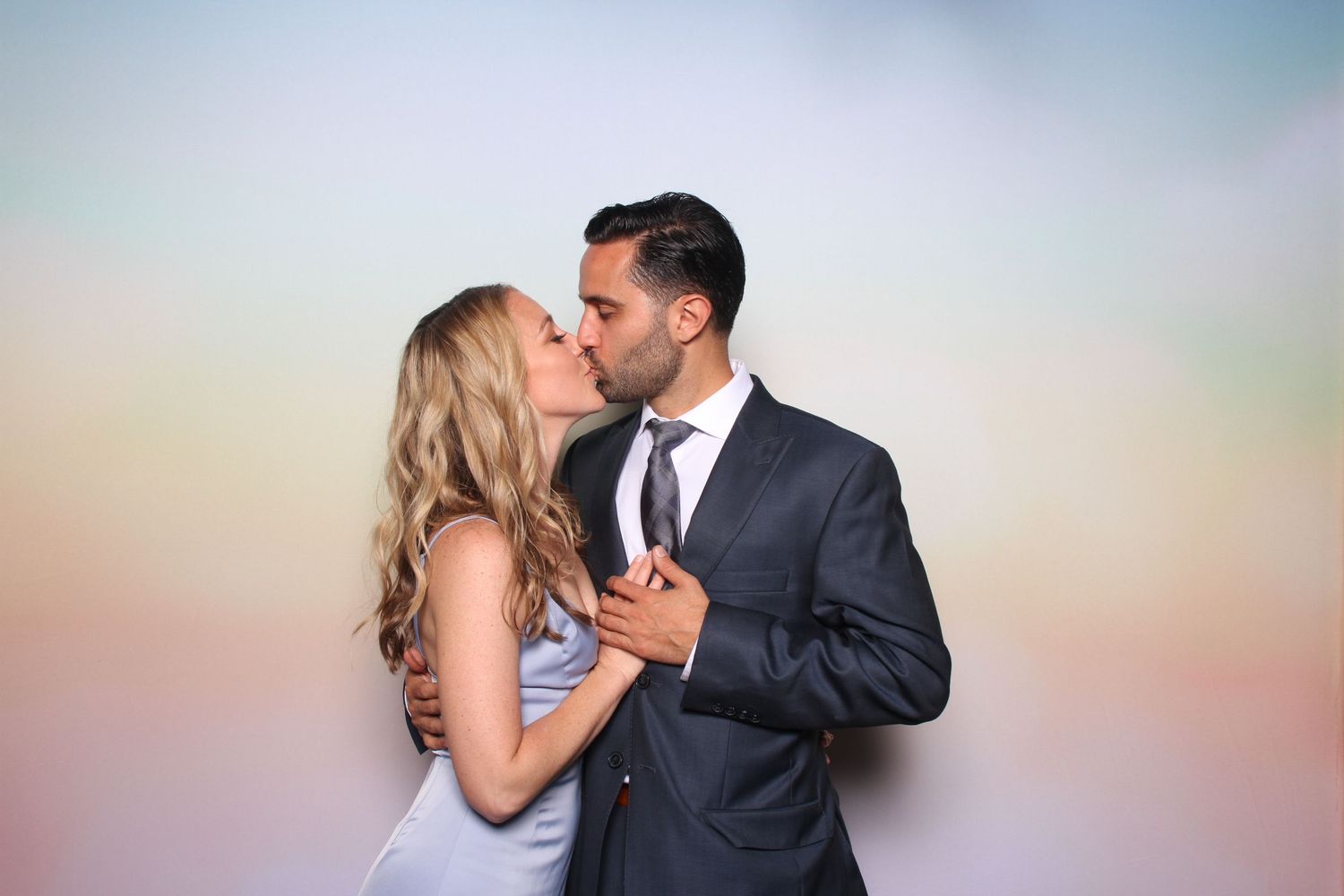 Couple sharing a romantic kiss at a photo booth with a soft pastel rainbow background.