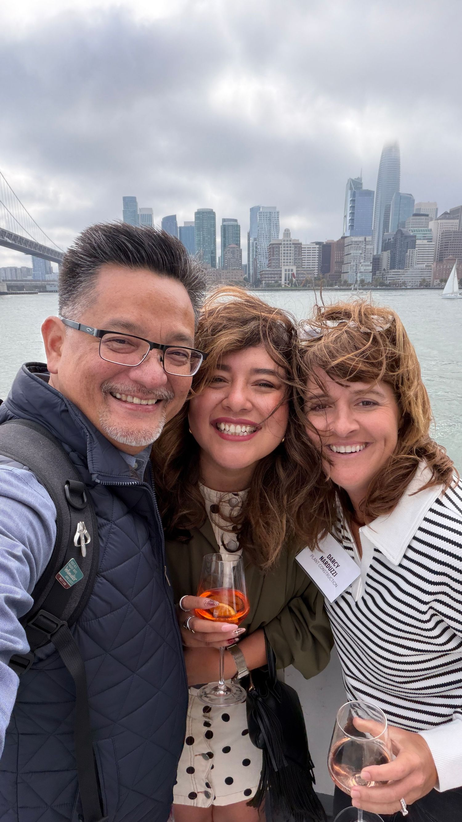 Three friends smiling for a selfie with wine glasses, San Francisco skyline and bay visible in the background.
